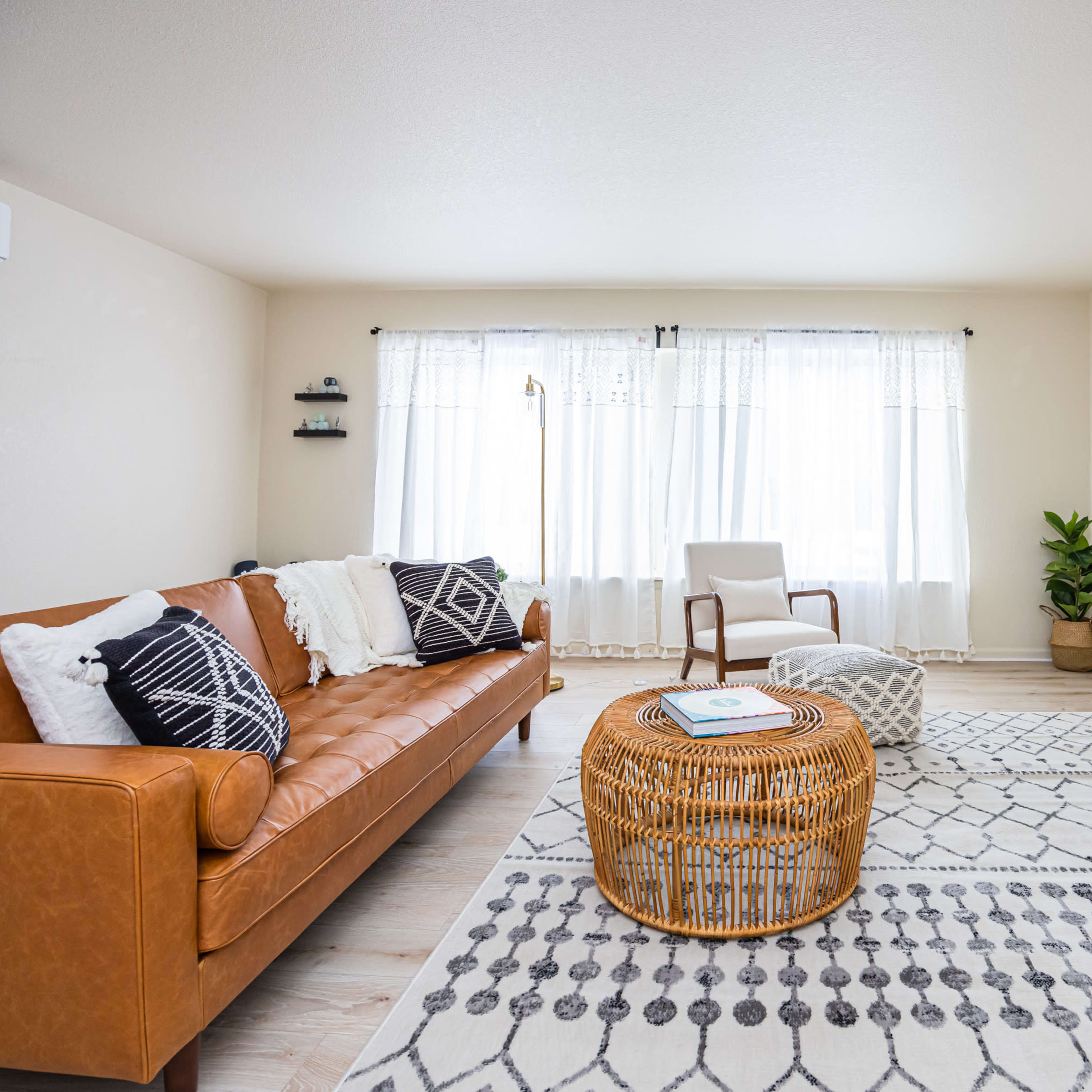 A bright and airy living room features a brown leather sofa, a round woven coffee table, and large windows with sheer curtains.
