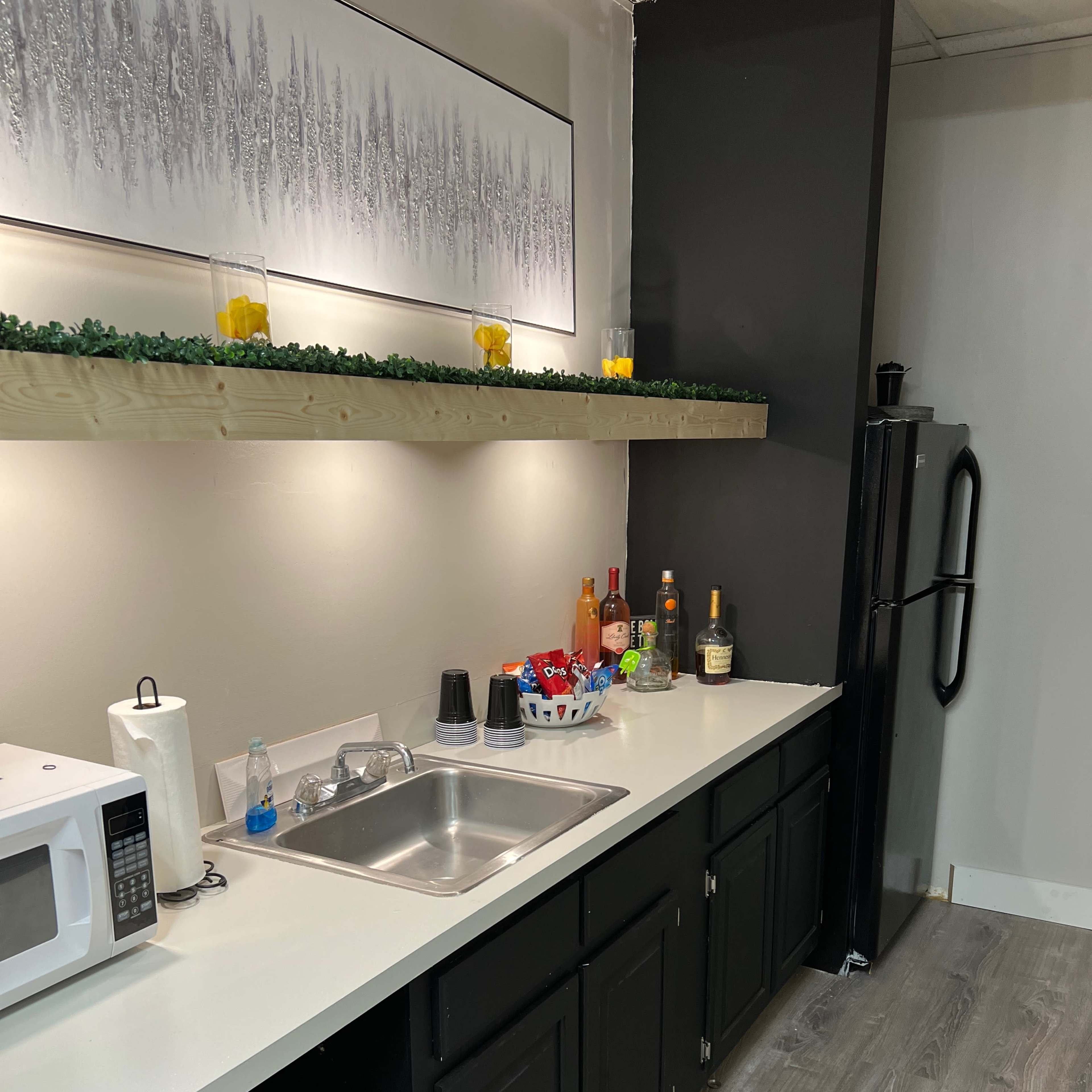 A kitchen with a black refrigerator, a stainless steel sink, a microwave, and a countertop shelf adorned with decorative items and multiple bottles of liquor.