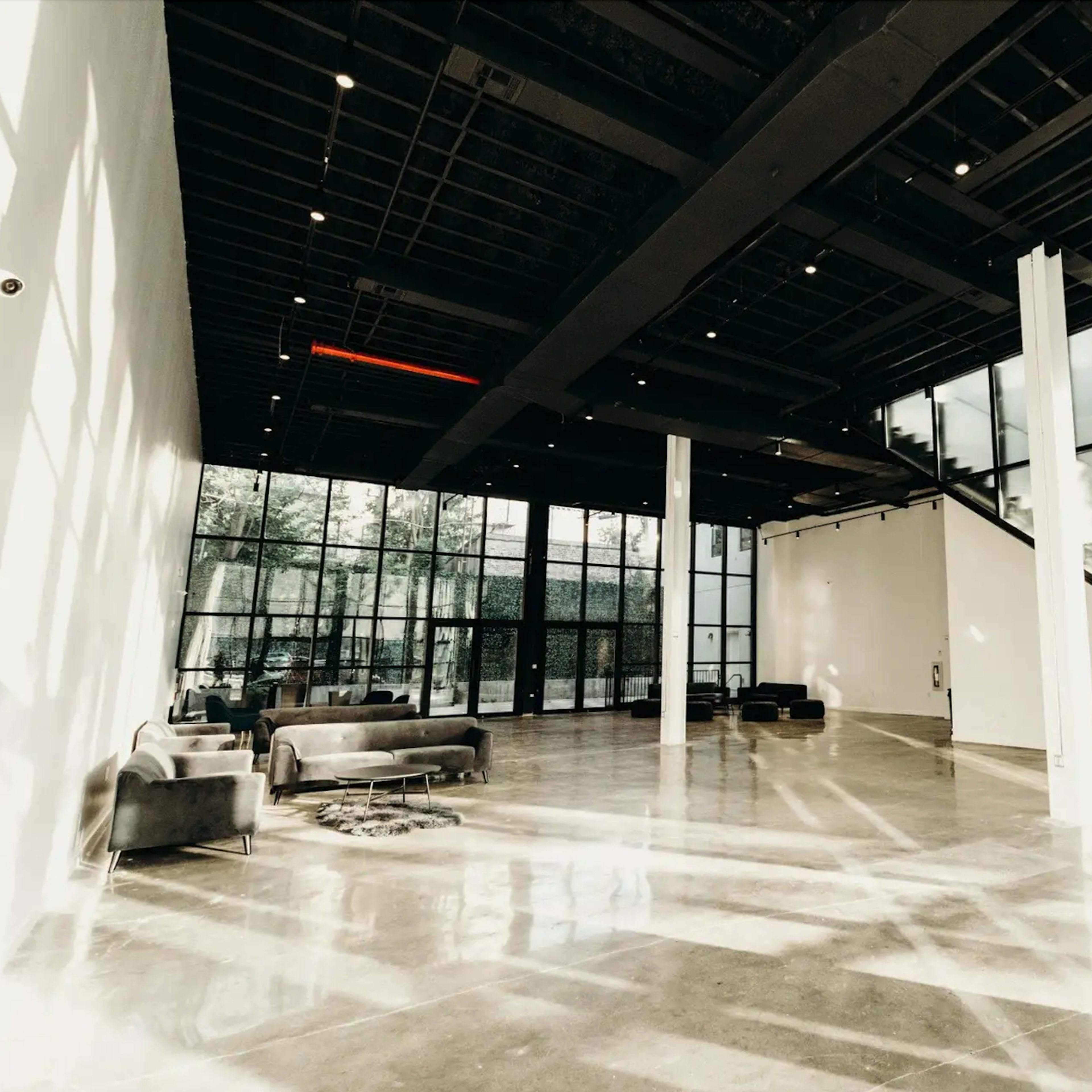 A spacious, modern interior featuring concrete floors, large glass windows, and minimalistic furniture arranged in an open area.