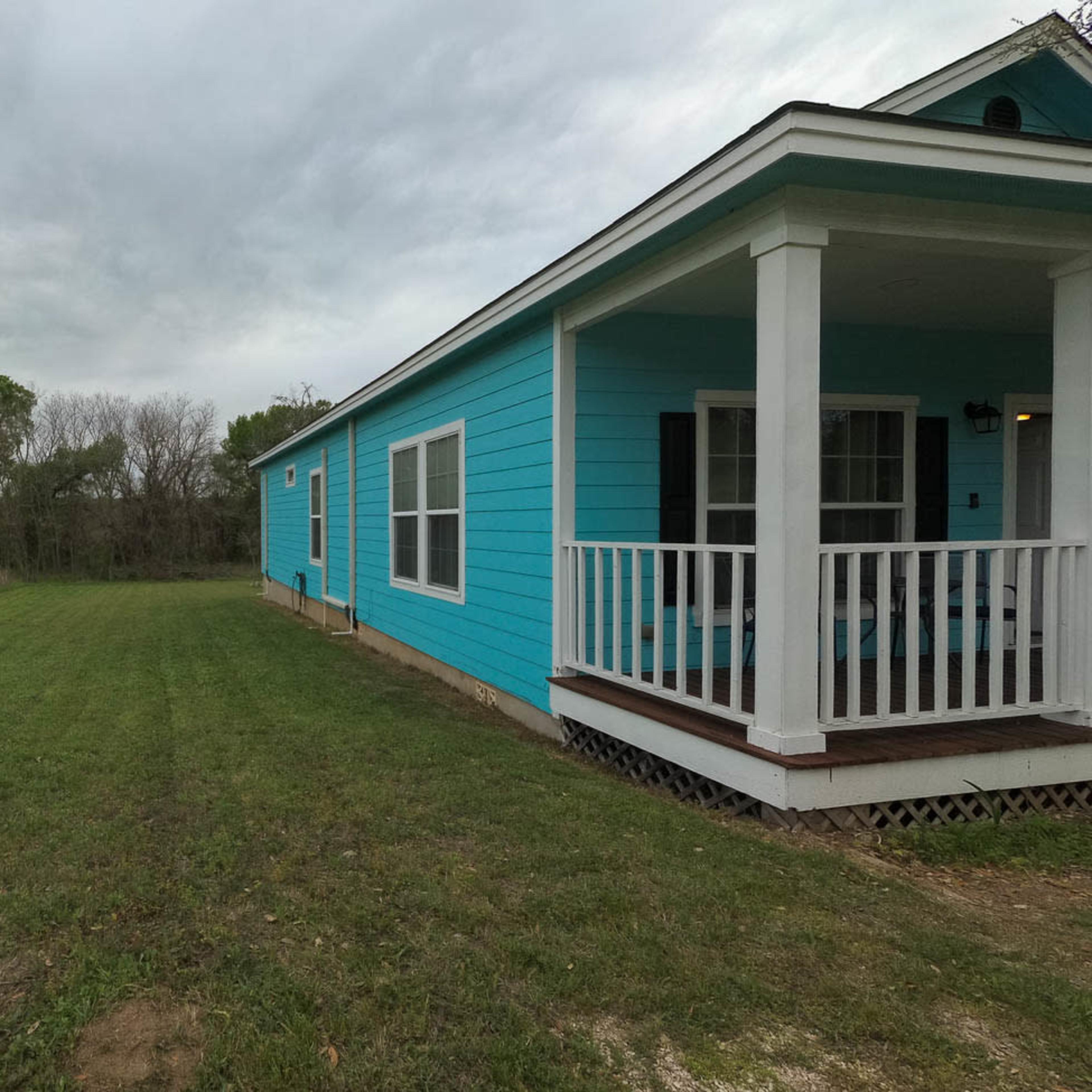 A blue house with a porch is situated next to a grassy field under a cloudy sky.