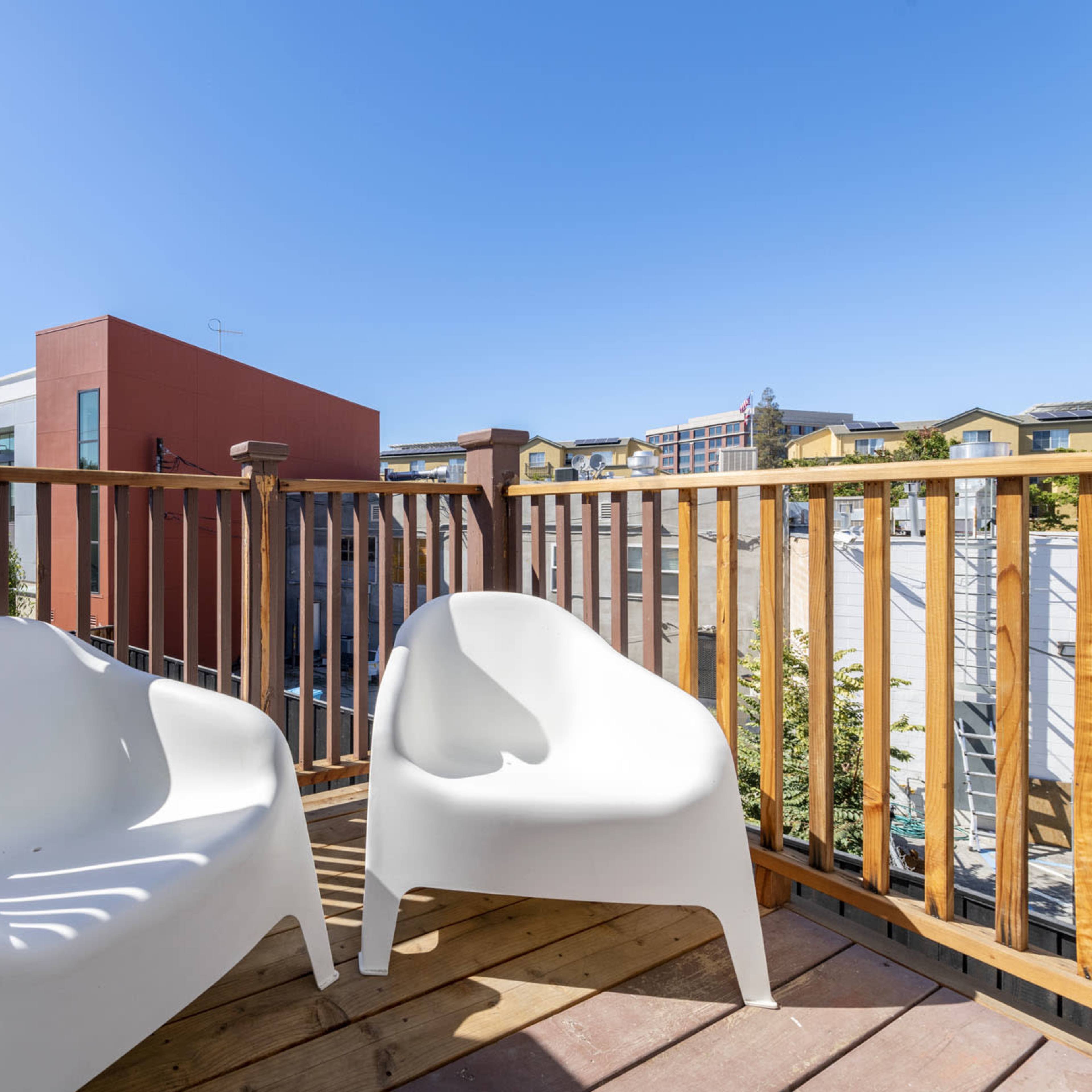 A small wooden balcony enclosed by a railing, featuring two white chairs and a clear blue sky overhead.