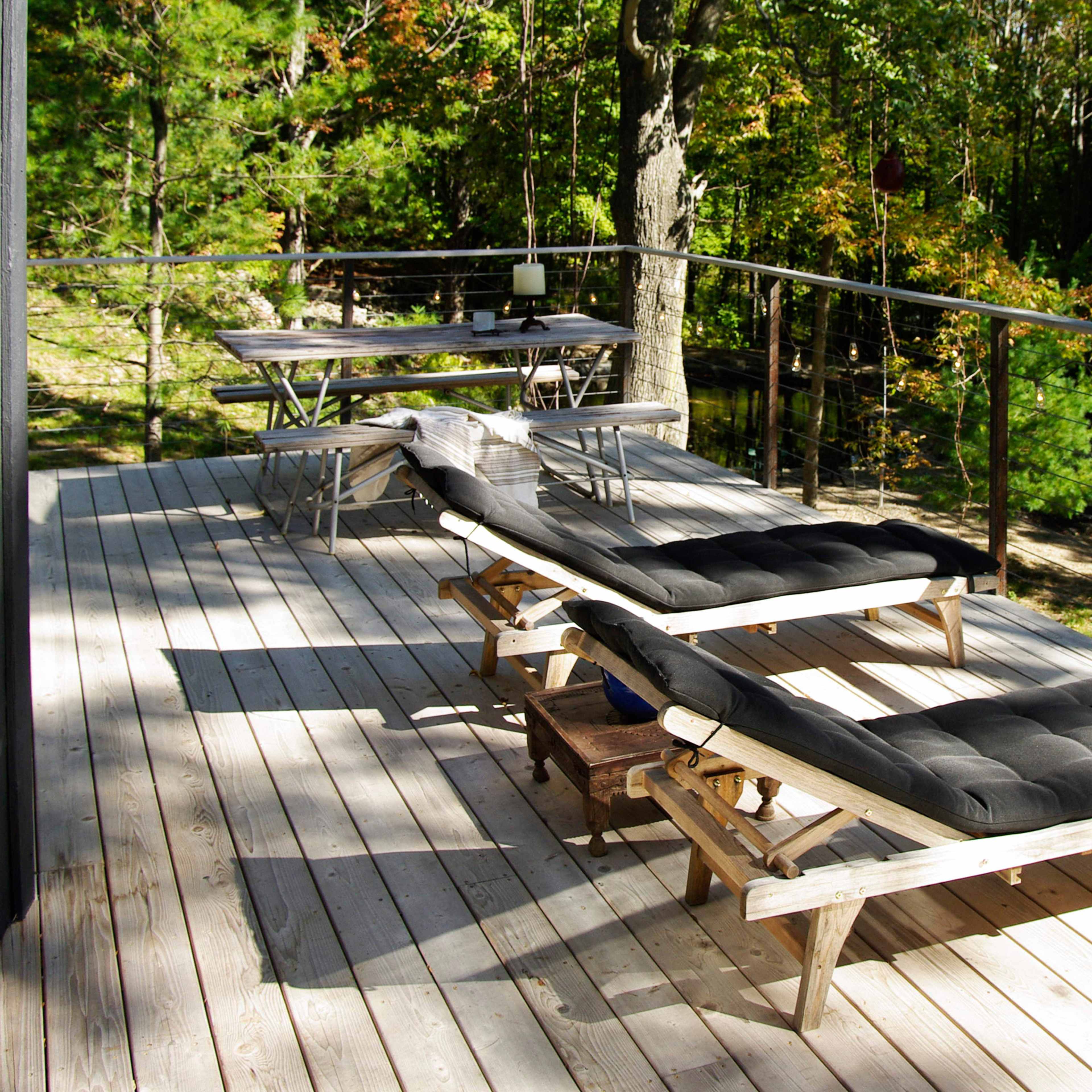 A wooden deck with two black lounge chairs and a table surrounded by trees.