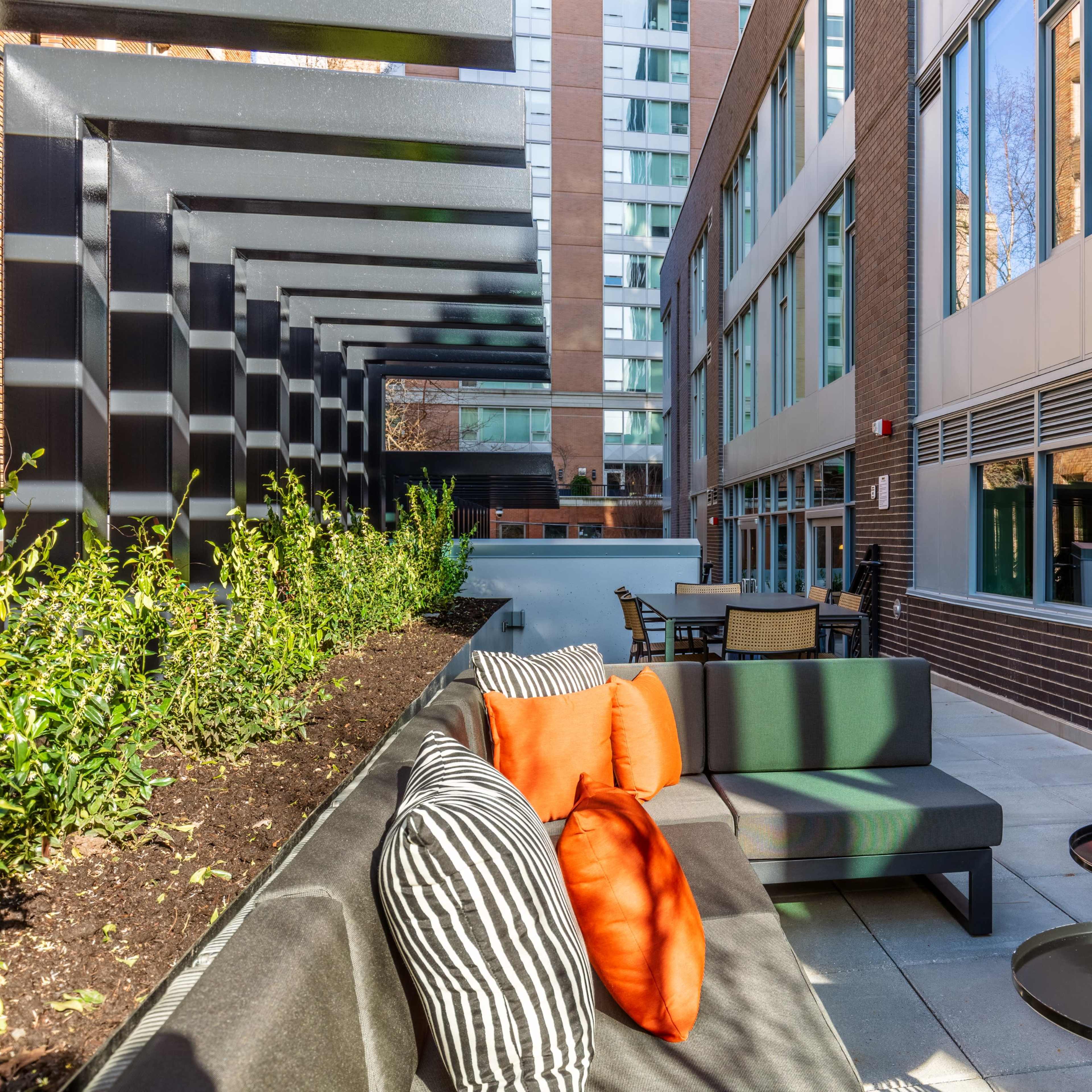 A modern outdoor seating area features a sectional couch with orange cushions, surrounded by greenery and brick buildings.