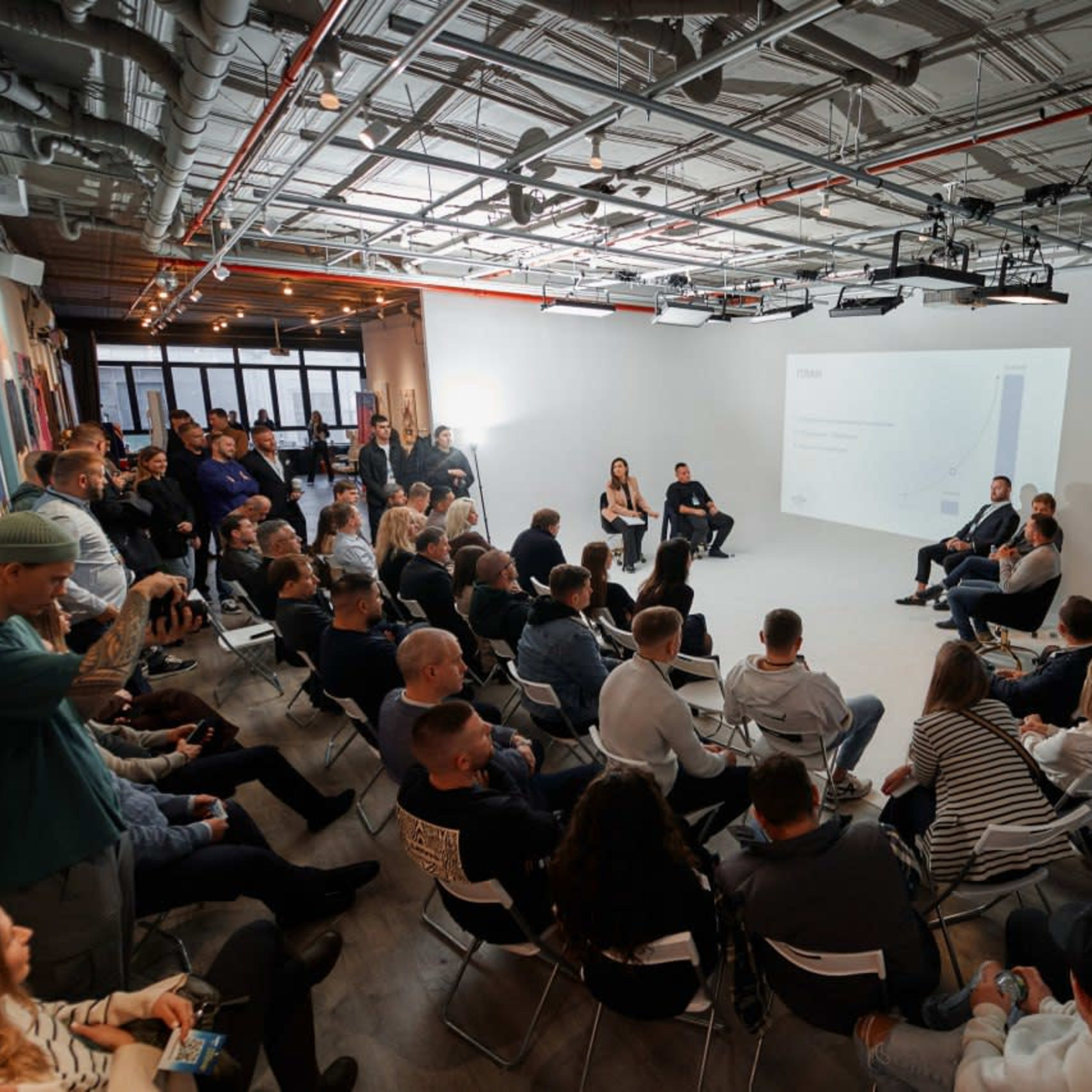 A crowd of people is seated in a modern, well-lit space for a panel discussion event.