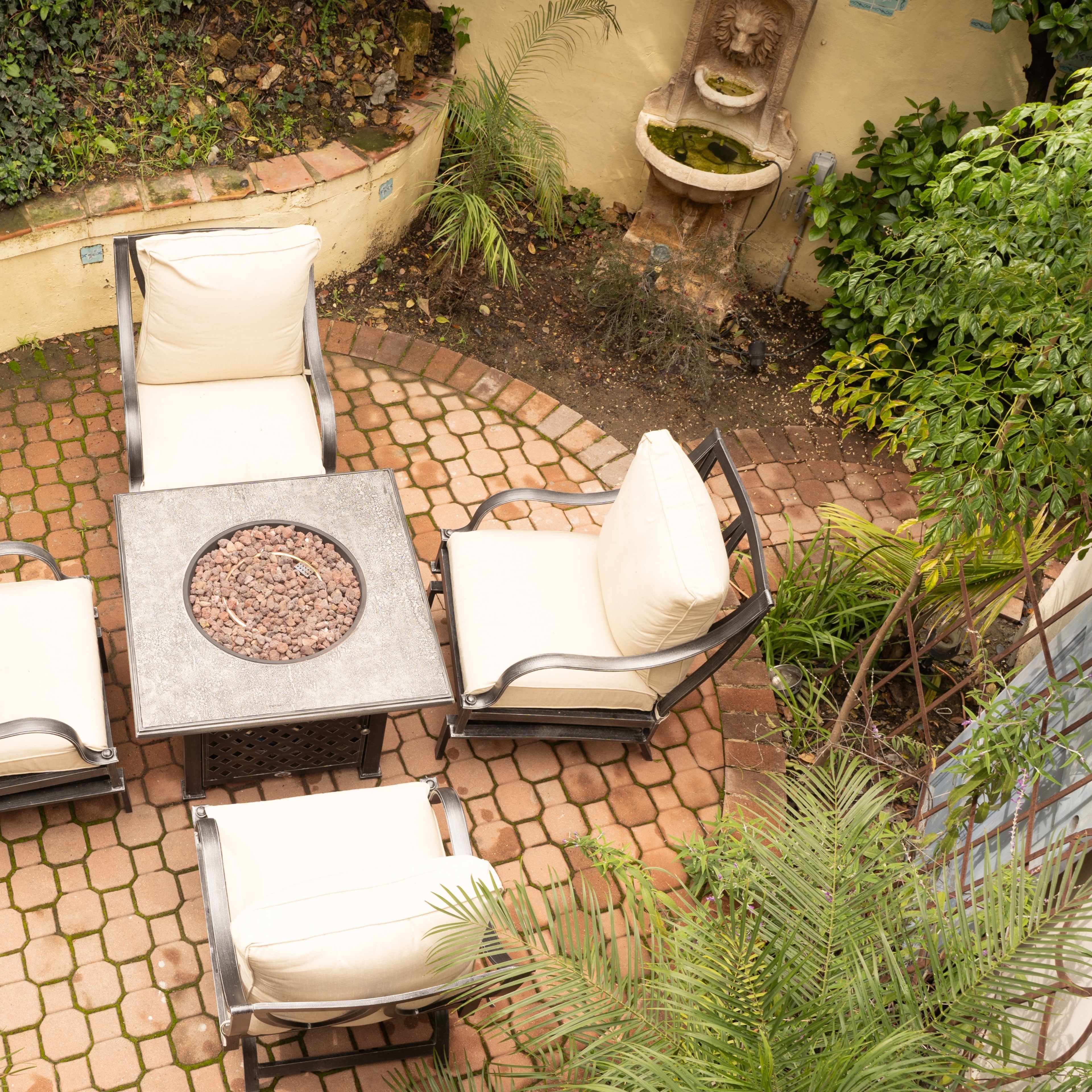 A circular seating area with four chairs and a central fire pit is surrounded by lush greenery and brick paving.