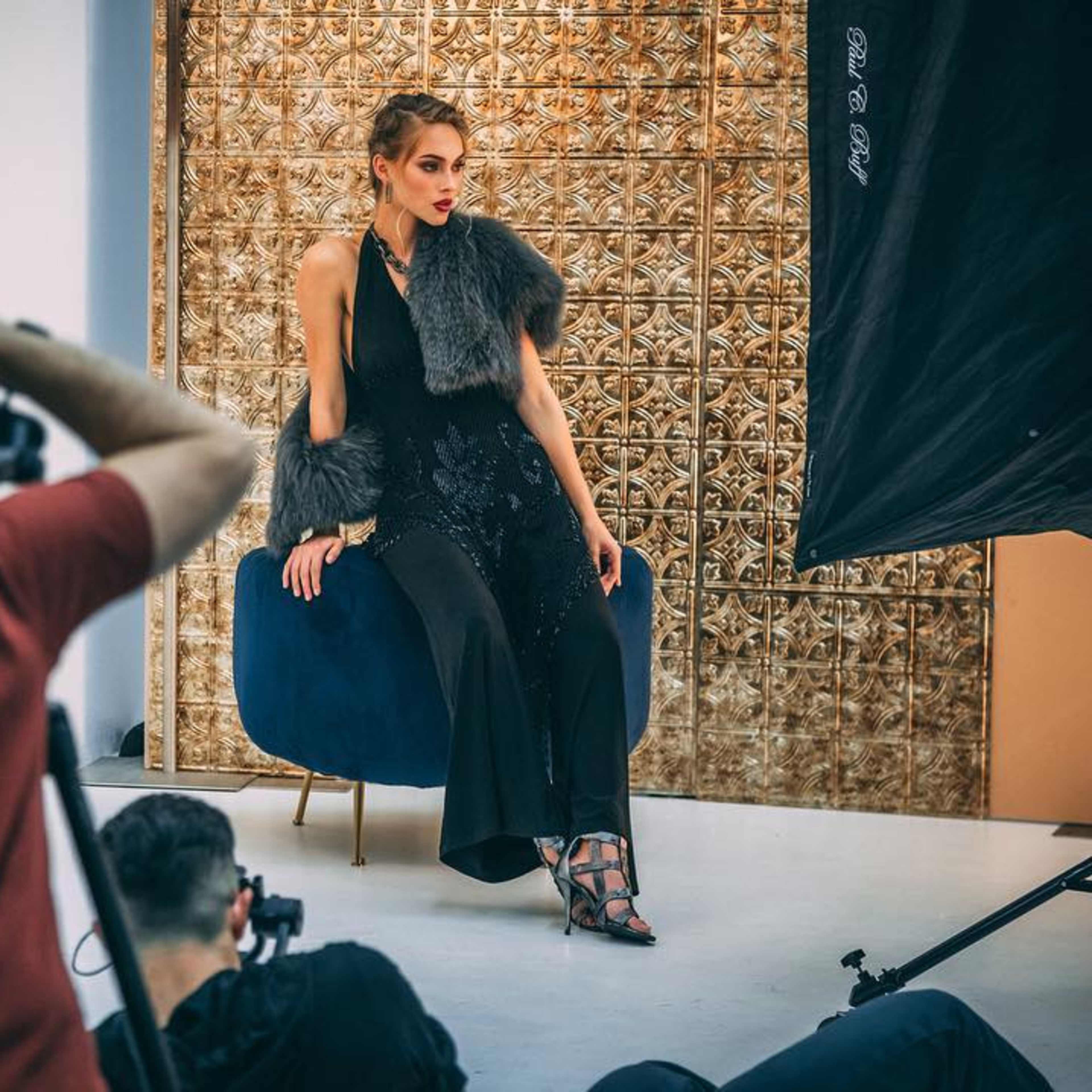 A model poses on a blue velvet chair against a textured gold backdrop while photographers capture the scene.