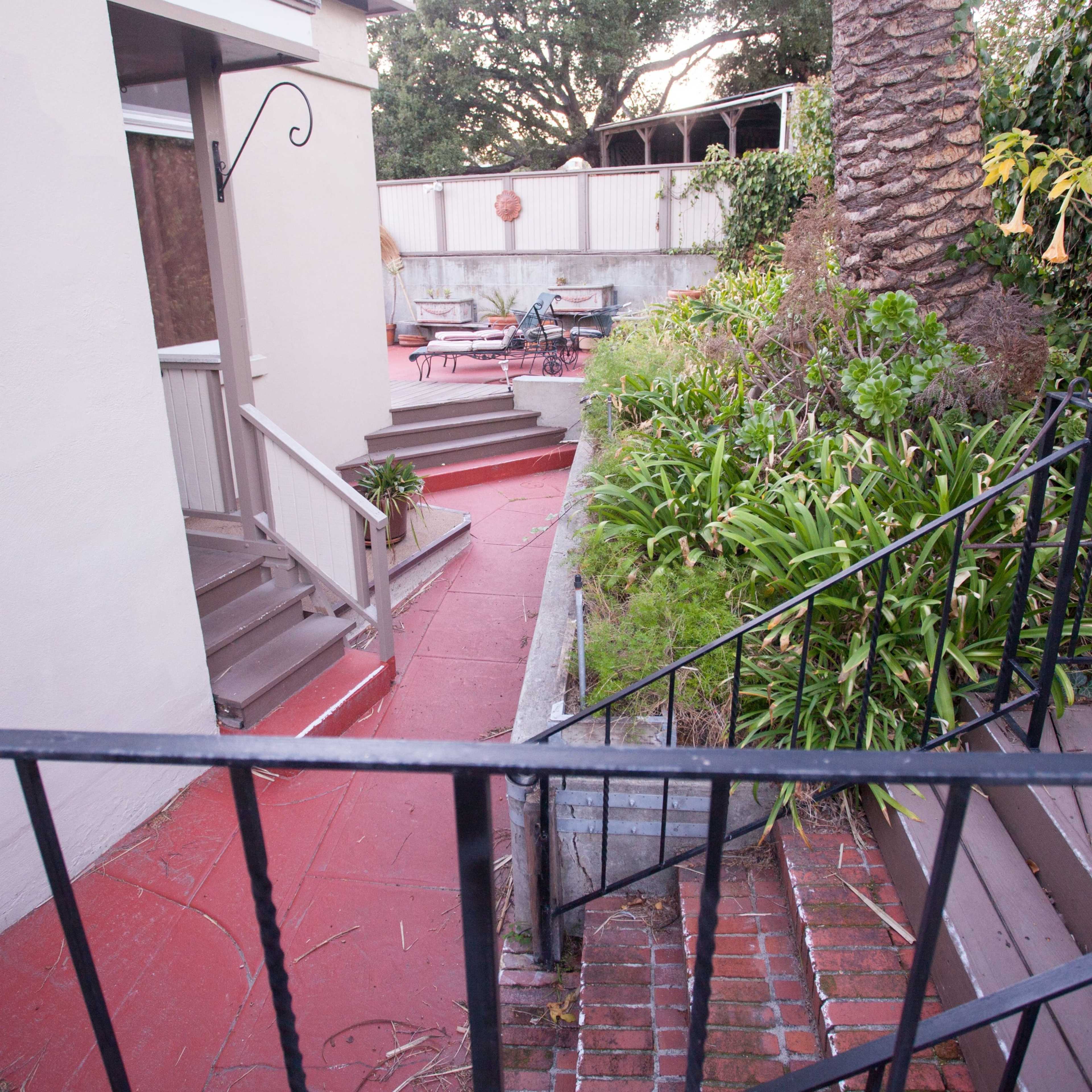 A pathway leading from a house to a garden area, with stairs on the right and a patio visible in the background.
