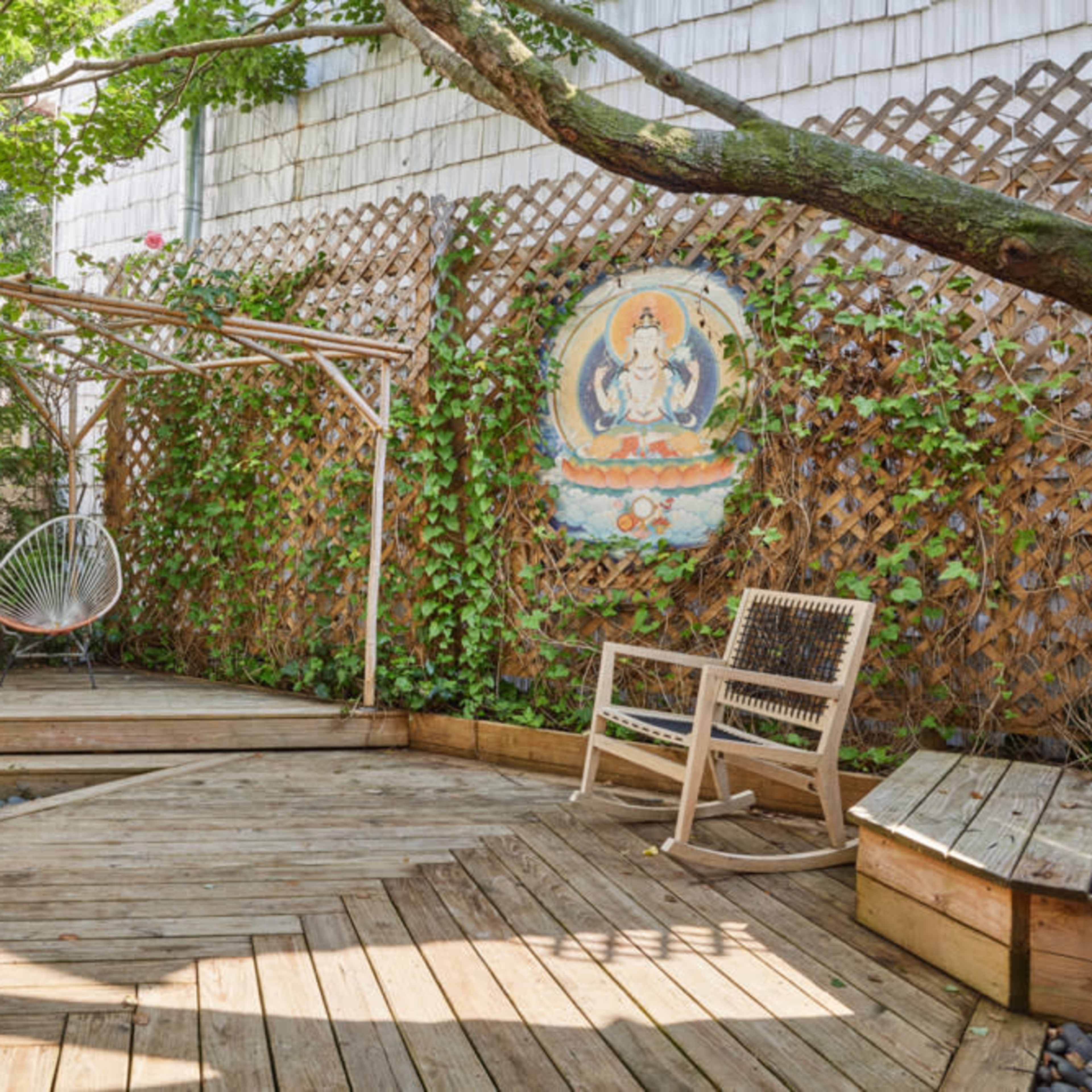 A wooden deck surrounded by greenery, featuring a hanging chair and a decorative wall with an ornate painting.