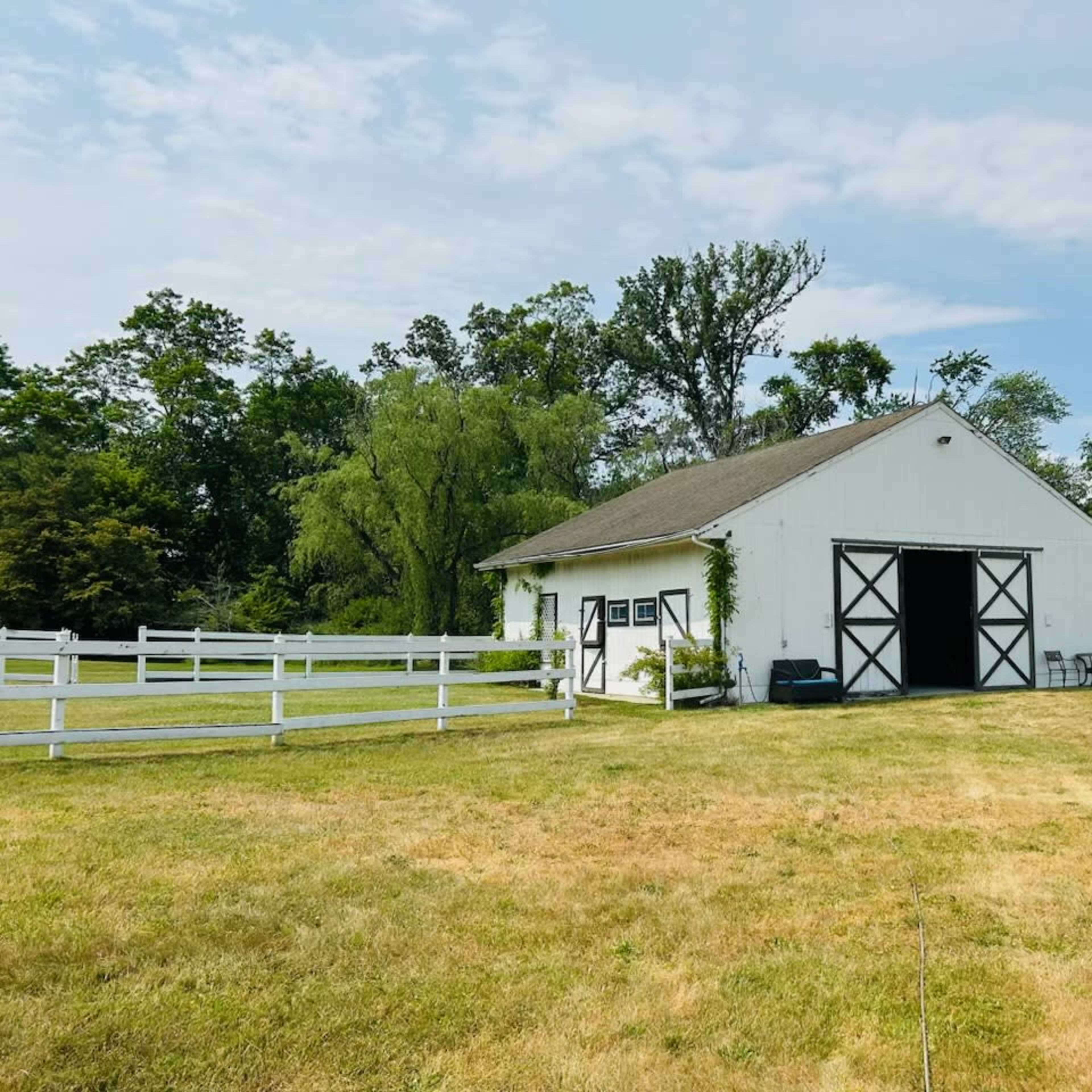 A white barn situated on a grassy field, surrounded by trees and a white fence.