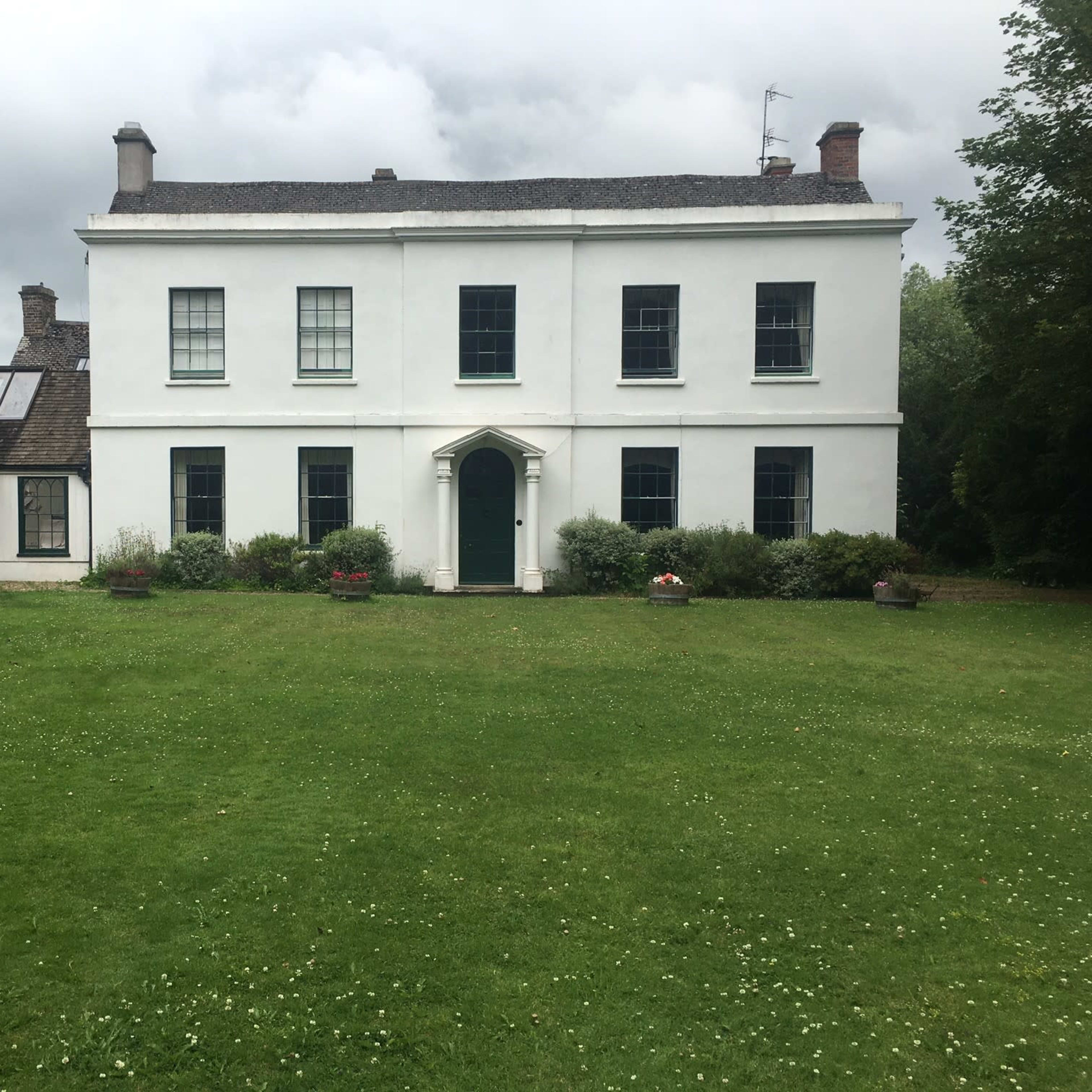 A white two-story house with green windows and a front door is situated on a grassy lawn surrounded by trees and shrubs.
