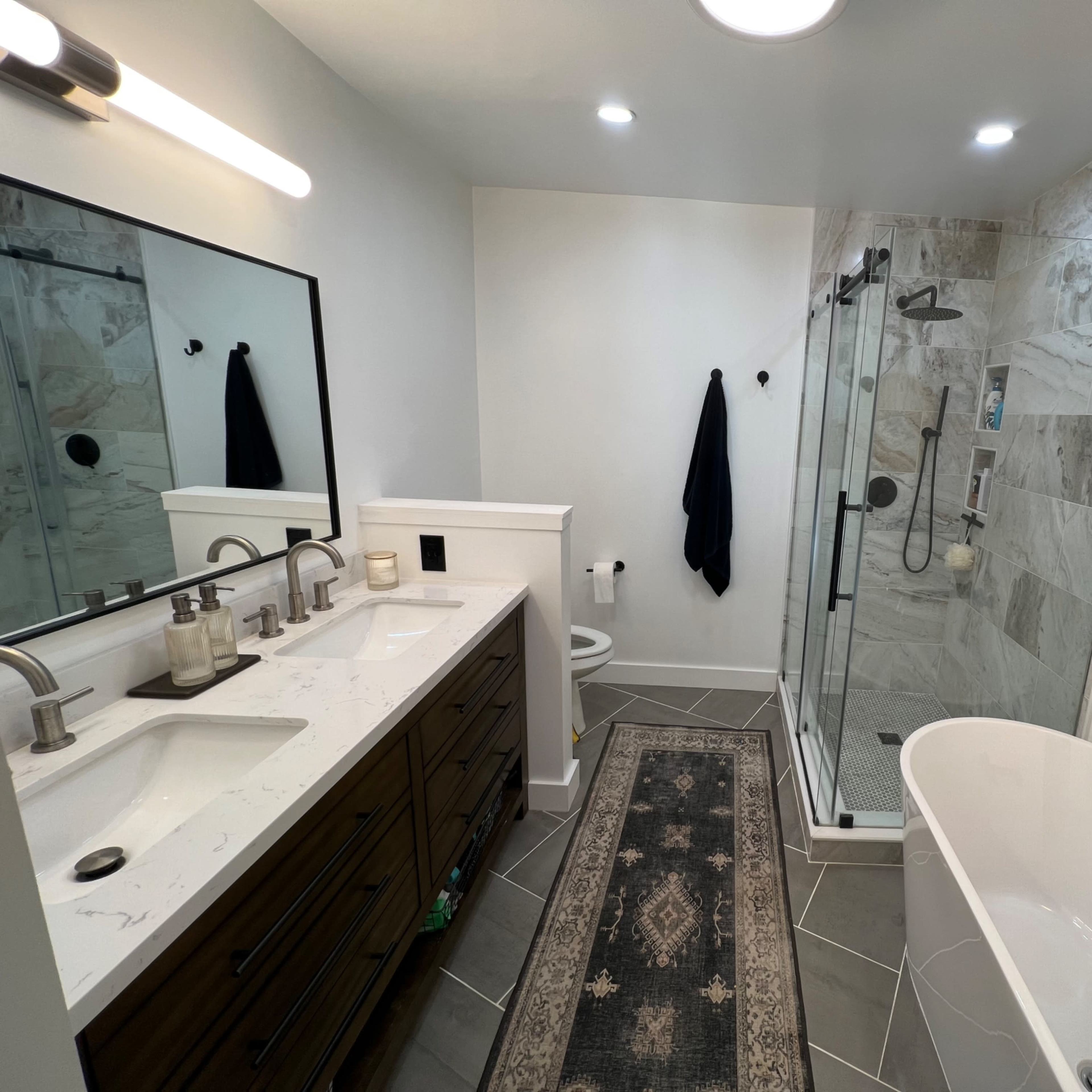 A modern bathroom featuring a double sink vanity, a walk-in shower, a freestanding bathtub, and a decorative rug on the tiled floor.