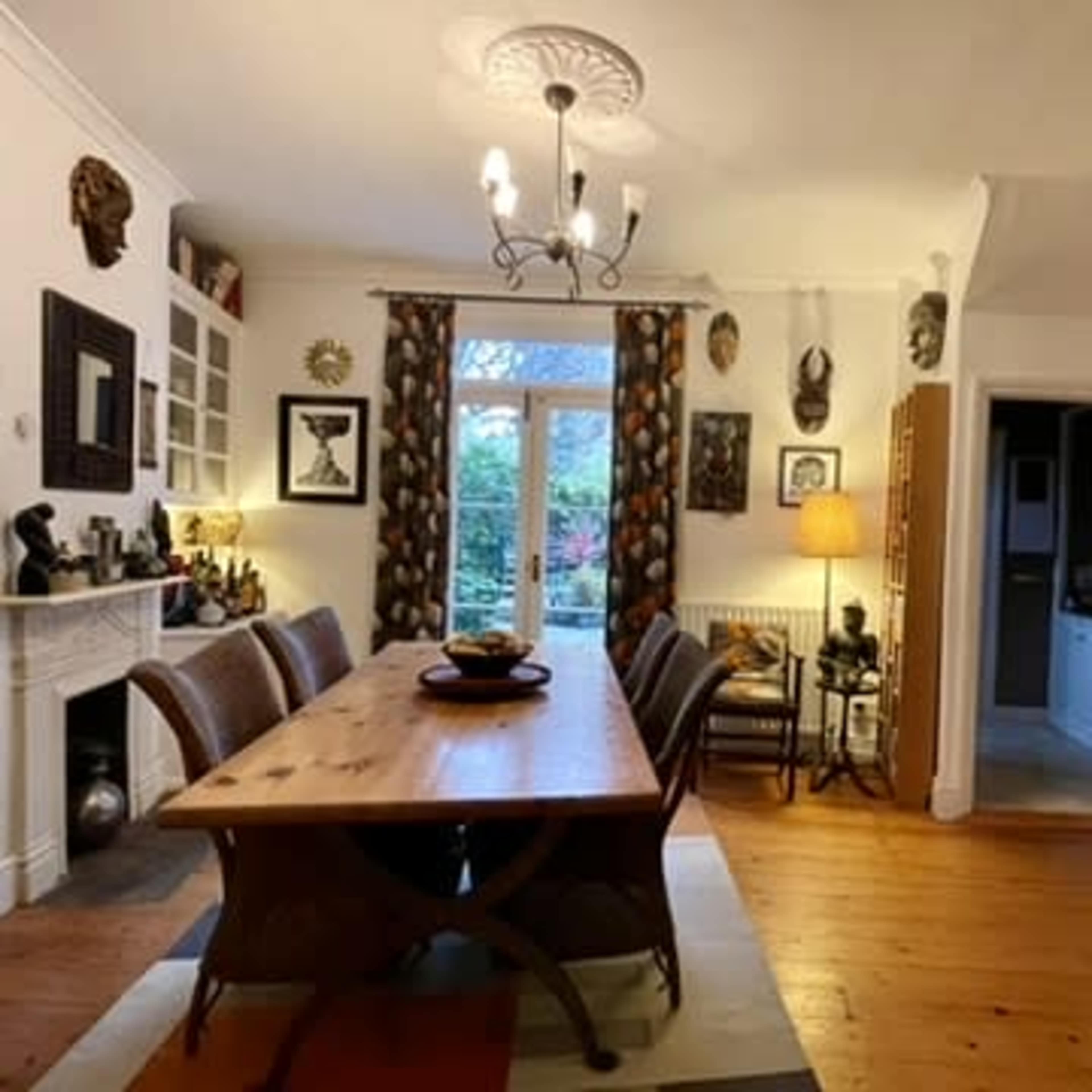A dining room features a long wooden table, surrounded by chairs, with a chandelier above and artwork and decorative masks on the walls.