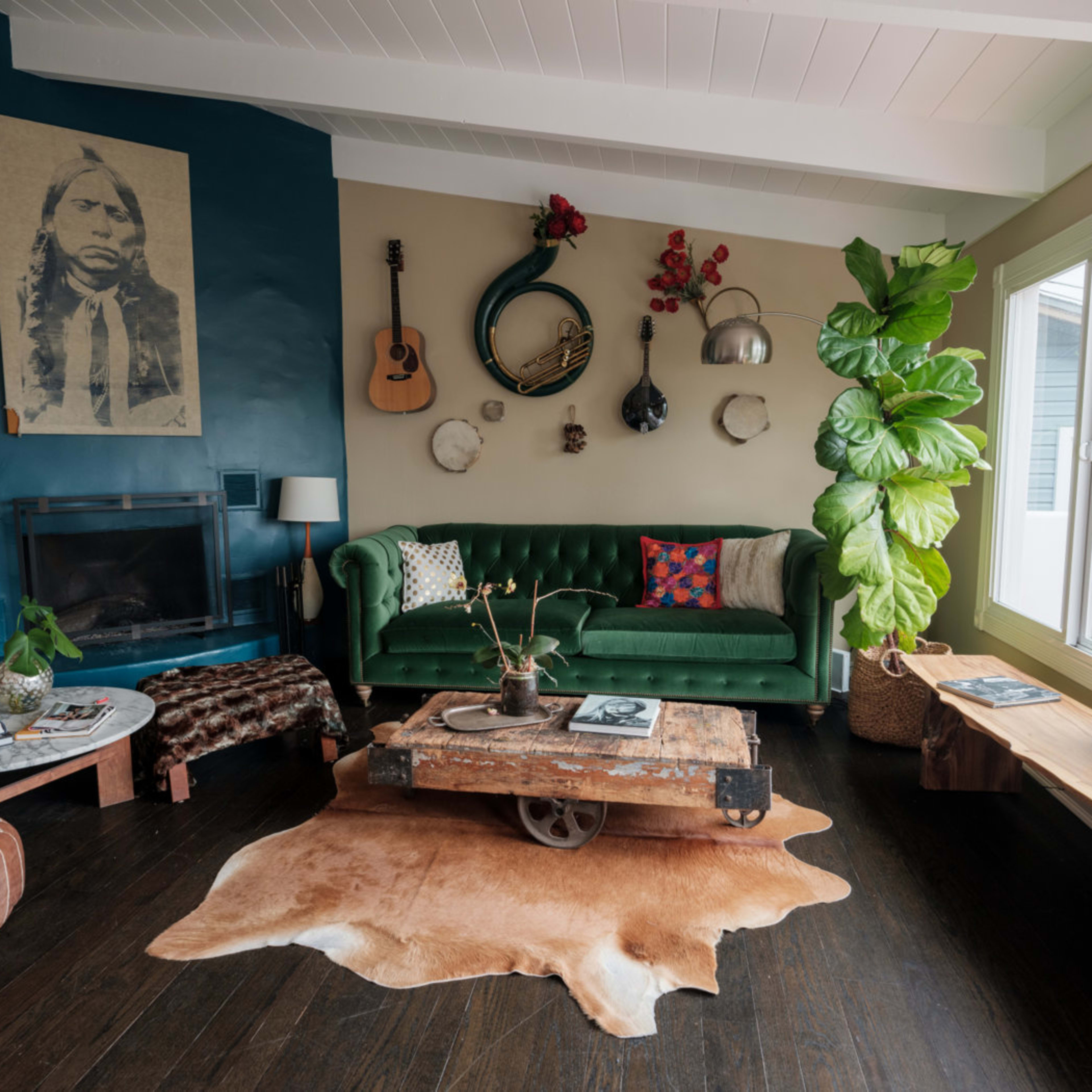 The room features a green sofa, a wooden coffee table on wheels, various musical instruments hanging on the wall, and large plants near a window.