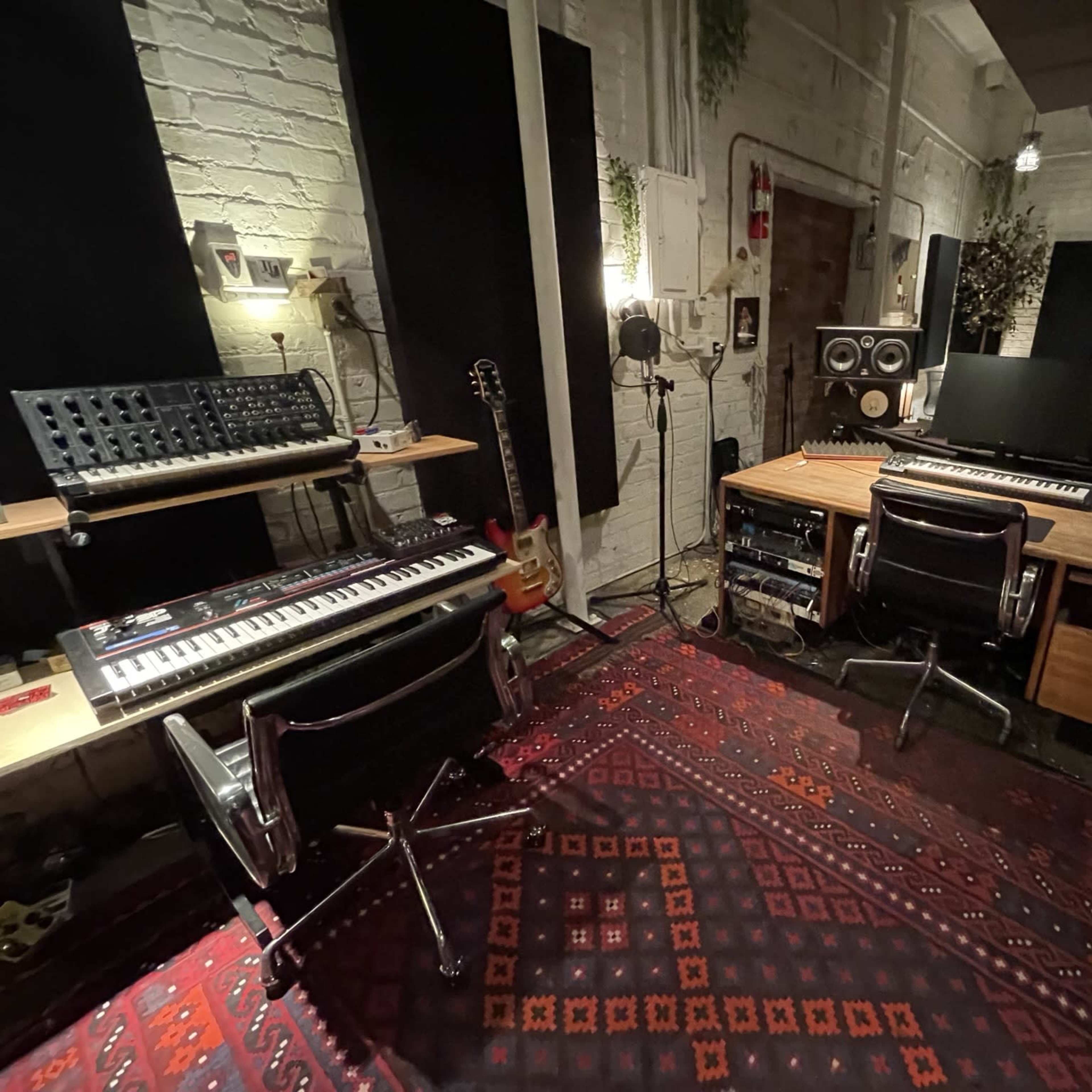 A music production studio with synthesizers, keyboards, and studio monitors positioned around a wooden desk on a patterned rug.