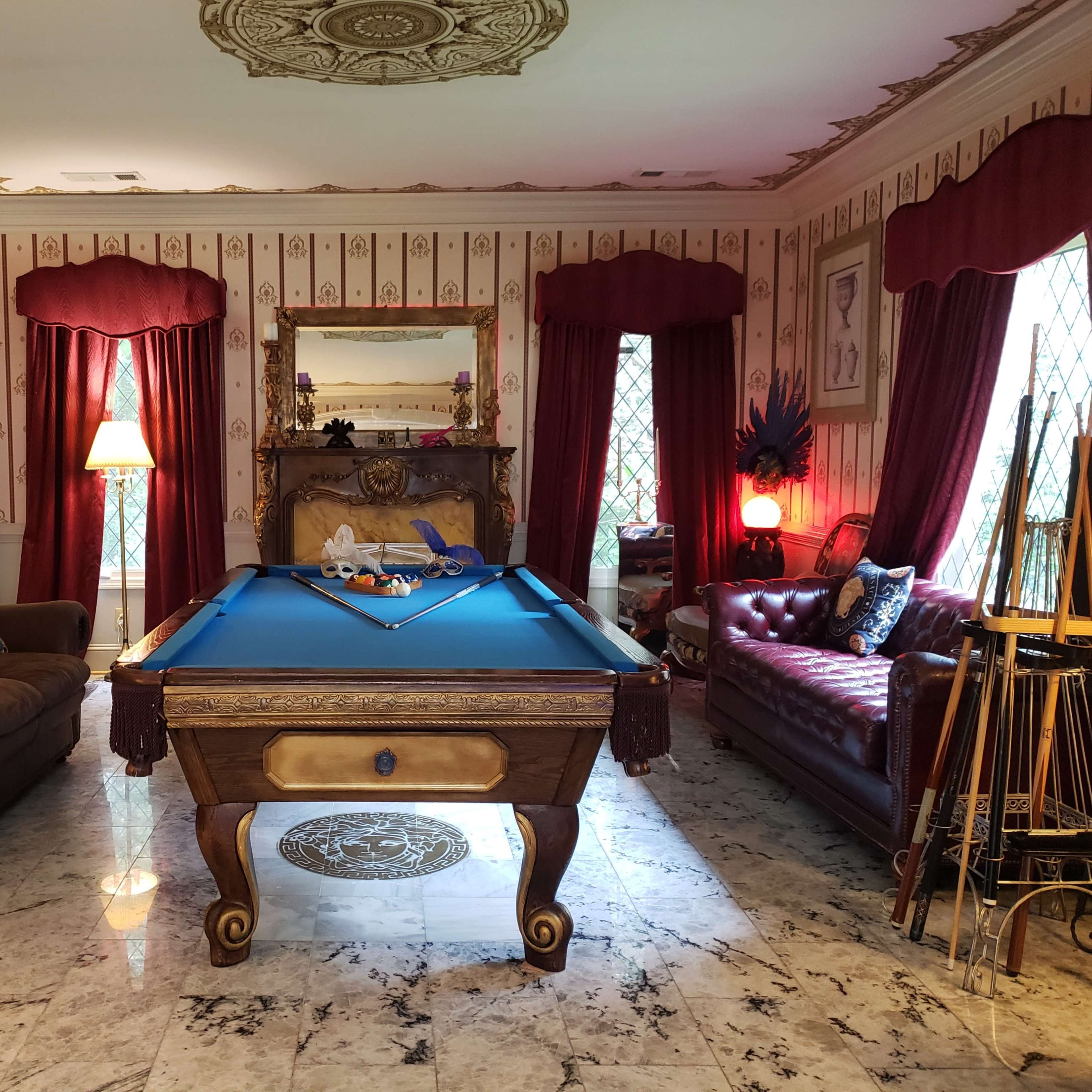 The room features a blue pool table at its center, surrounded by a leather couch, a patterned armchair, and red drapes framing two windows.