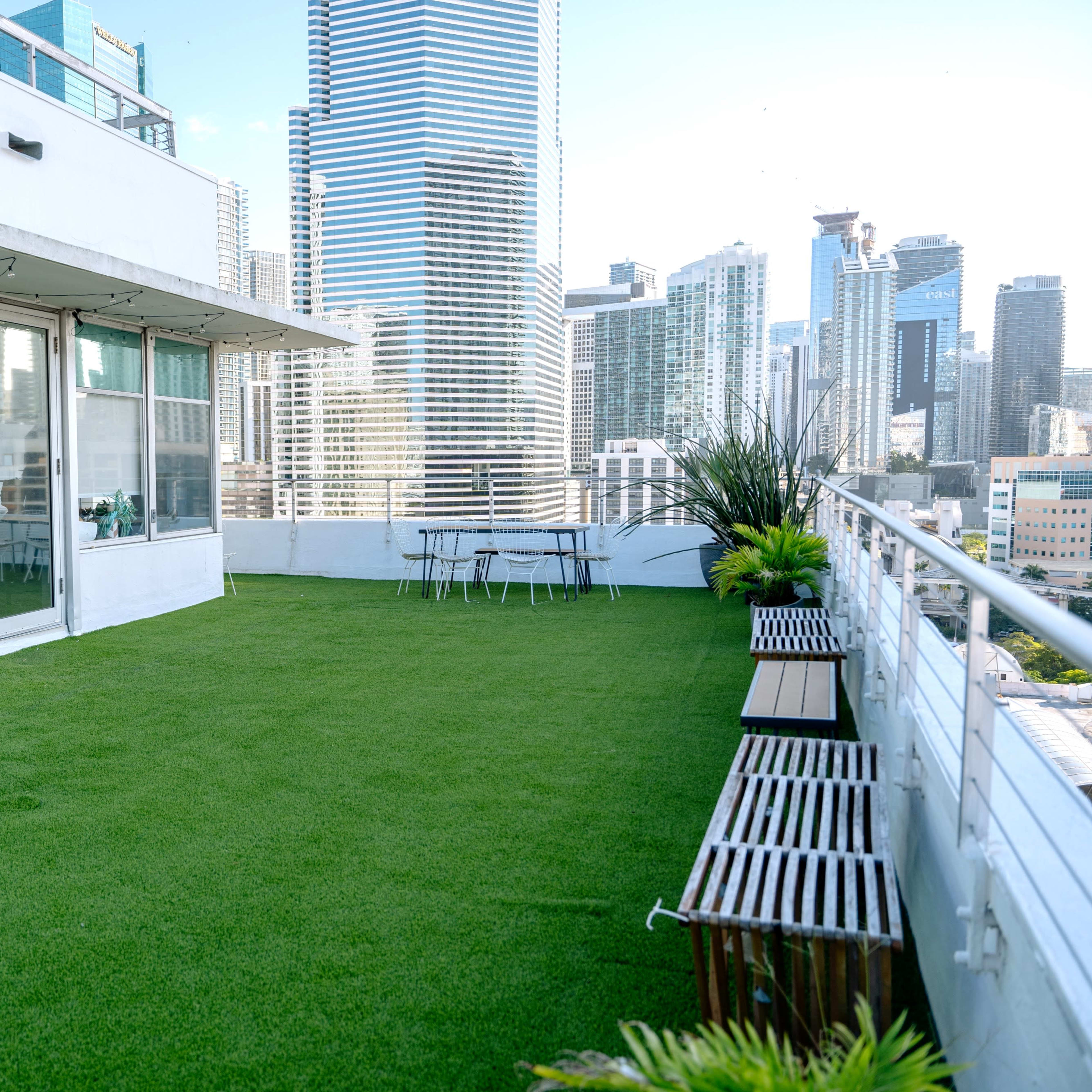 A spacious rooftop terrace features artificial grass, a table with chairs, and a view of tall city buildings.