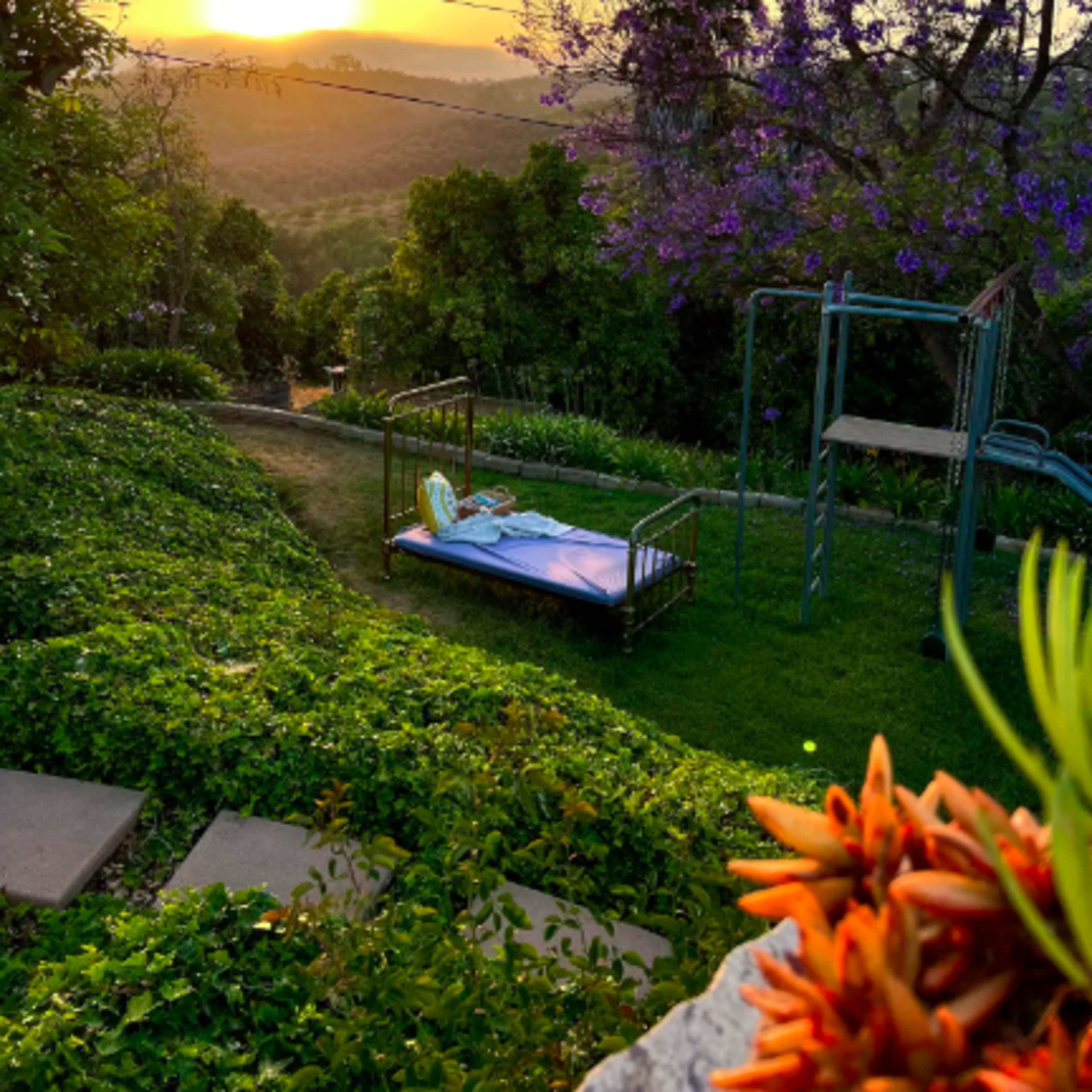 A bed is placed in a grassy area with a view of a sunset over distant hills, next to a playground swing set and colorful plants.
