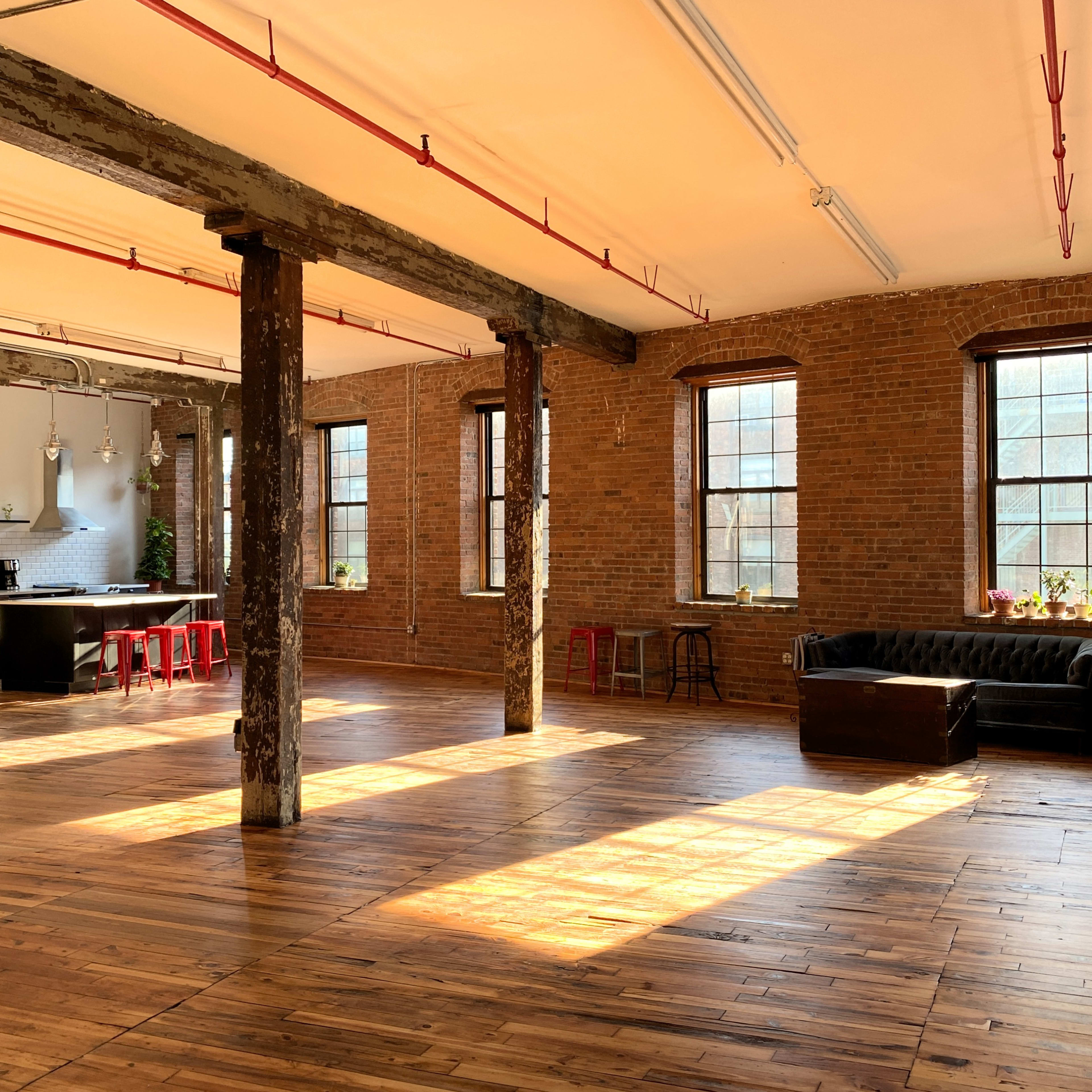 A spacious loft with exposed brick walls, wooden floors, and large windows, featuring a kitchen area, a leather sofa, and several potted plants.