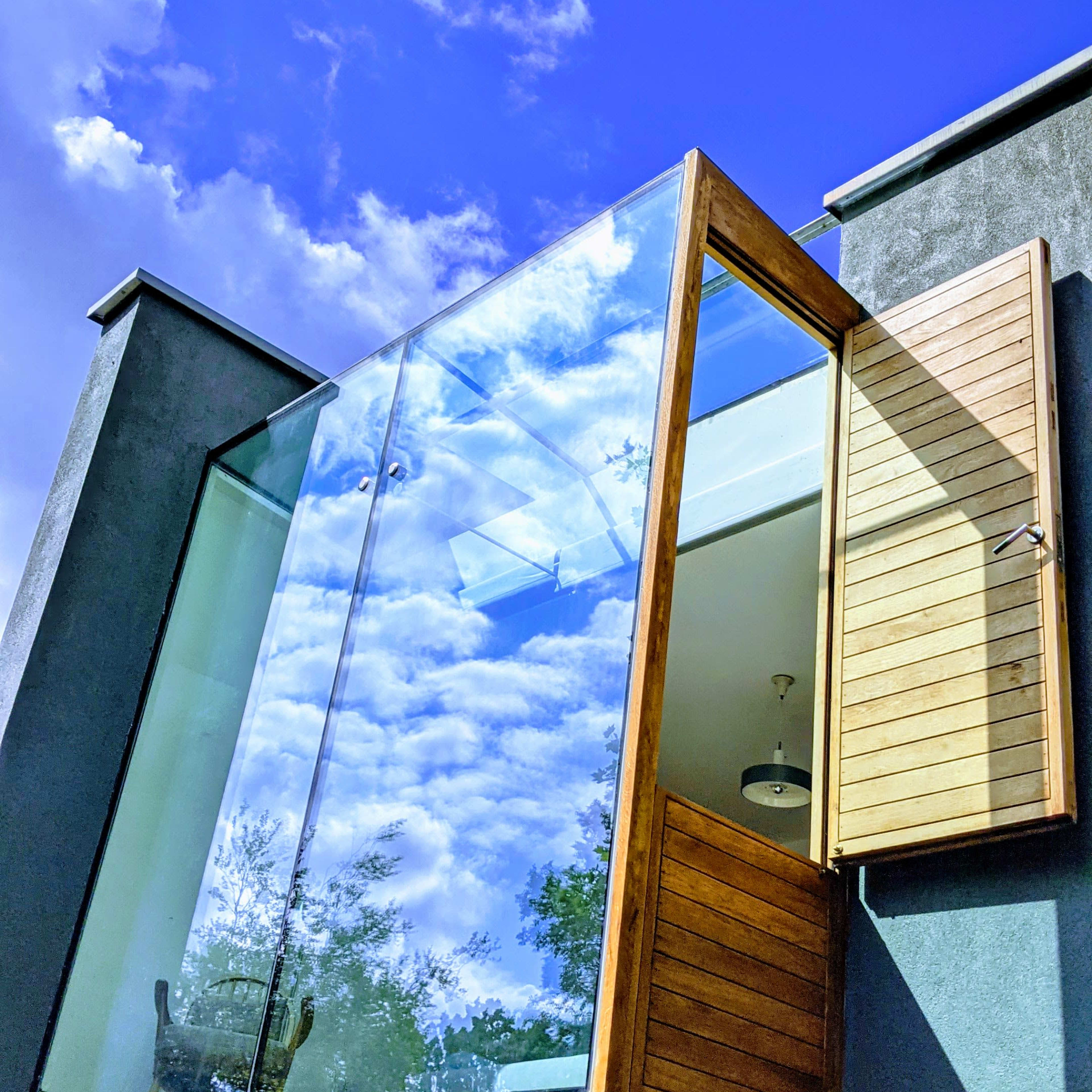 A modern balcony with a glass railing and wooden shutters is shown against a blue sky with scattered clouds.