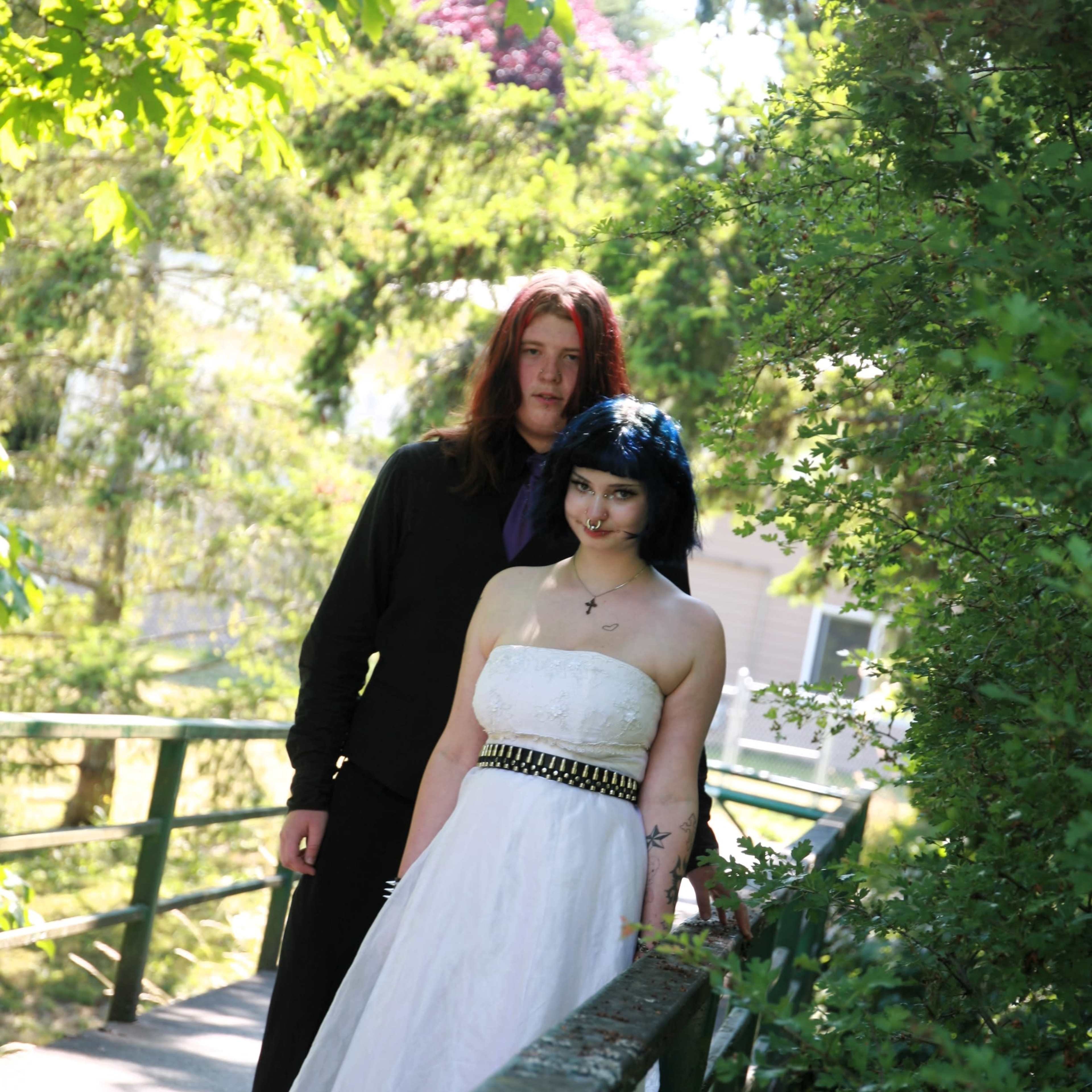 A man in a black outfit stands beside a woman in a white dress on a sunlit path surrounded by greenery.