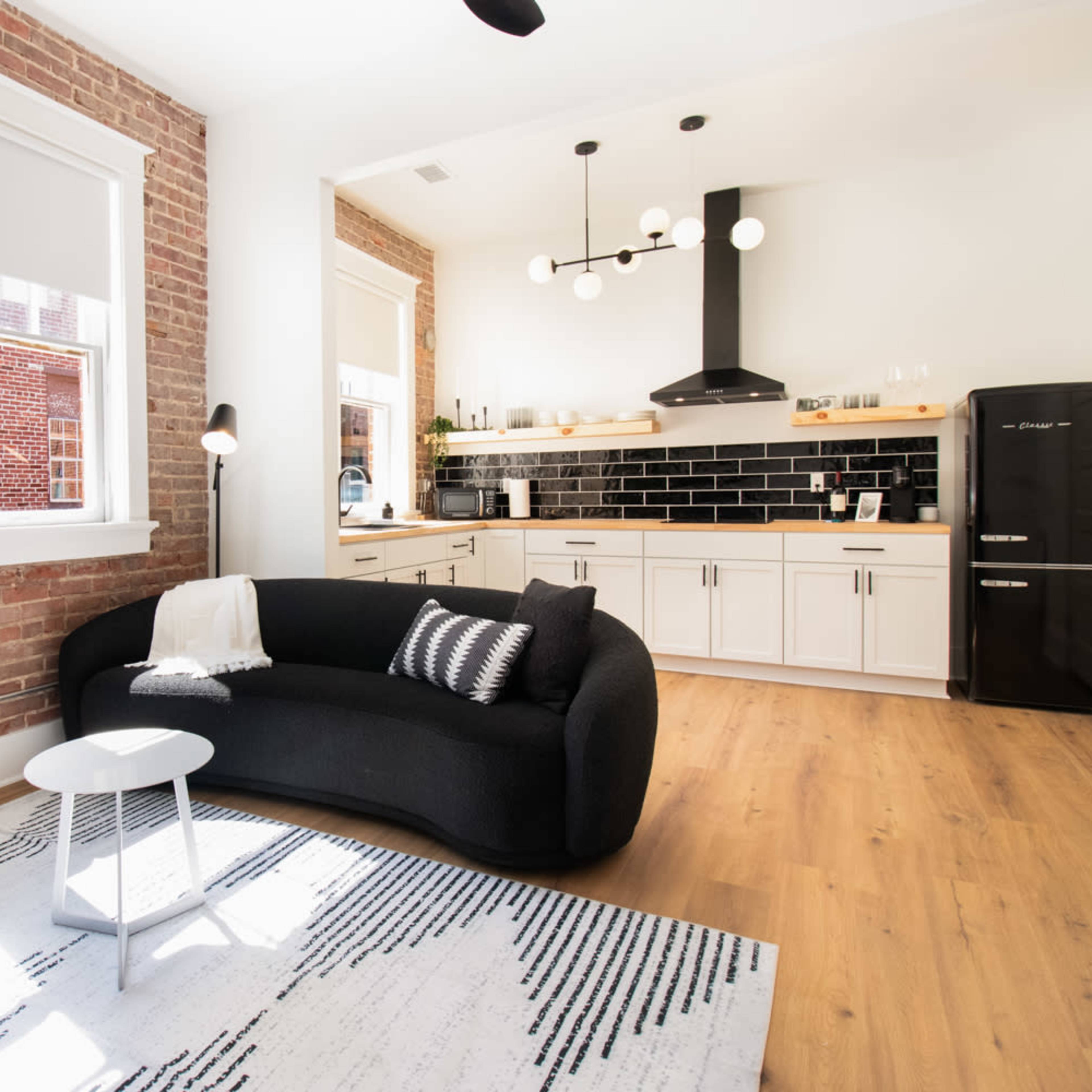 A modern living space featuring a black curved sofa, a round white side table, and a kitchen area with white cabinets and a black backsplash.