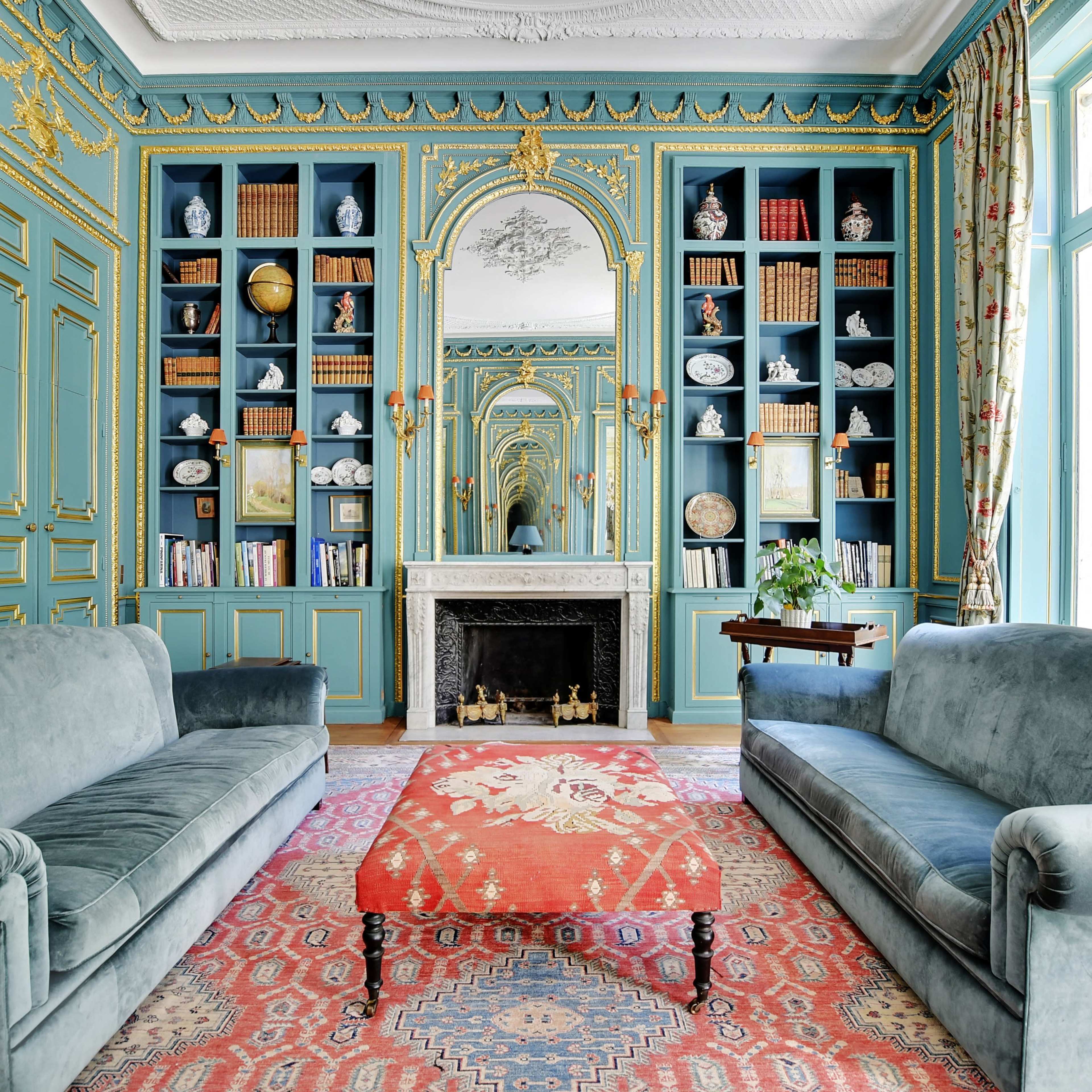 The room features two velvet sofas facing a central ornate rug, with a large bookshelf and a decorative fireplace against the blue walls.