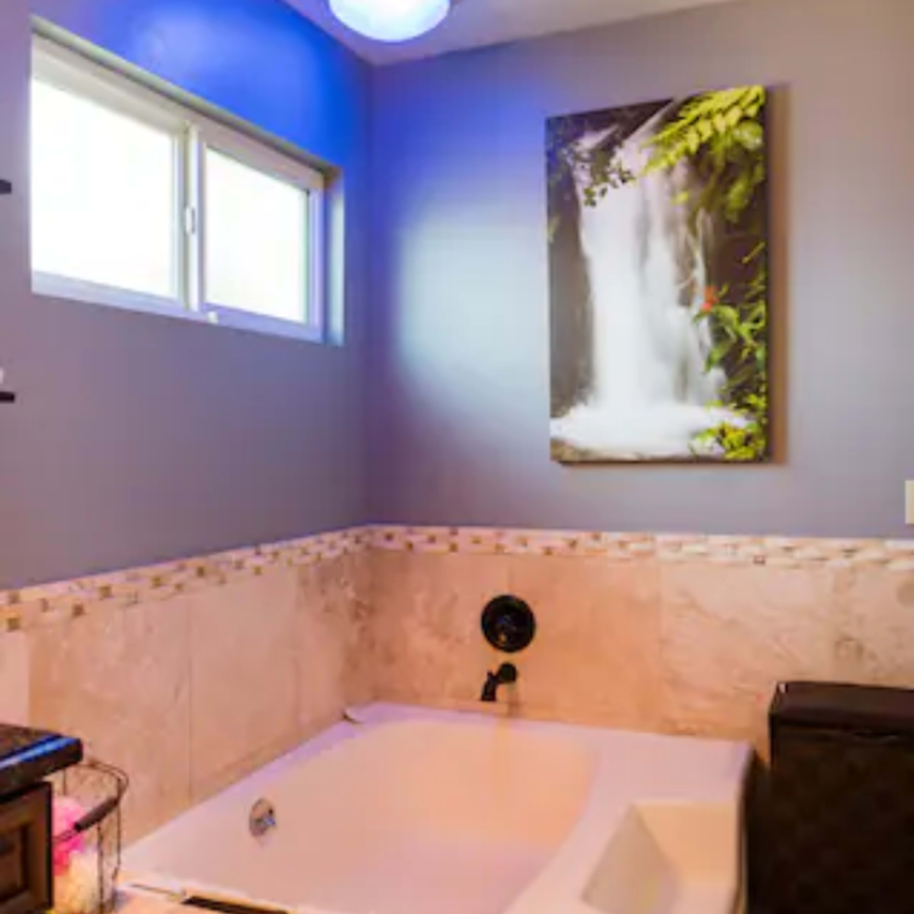 A bathroom featuring a jetted bathtub, a framed waterfall painting on the wall, and a window allowing natural light.