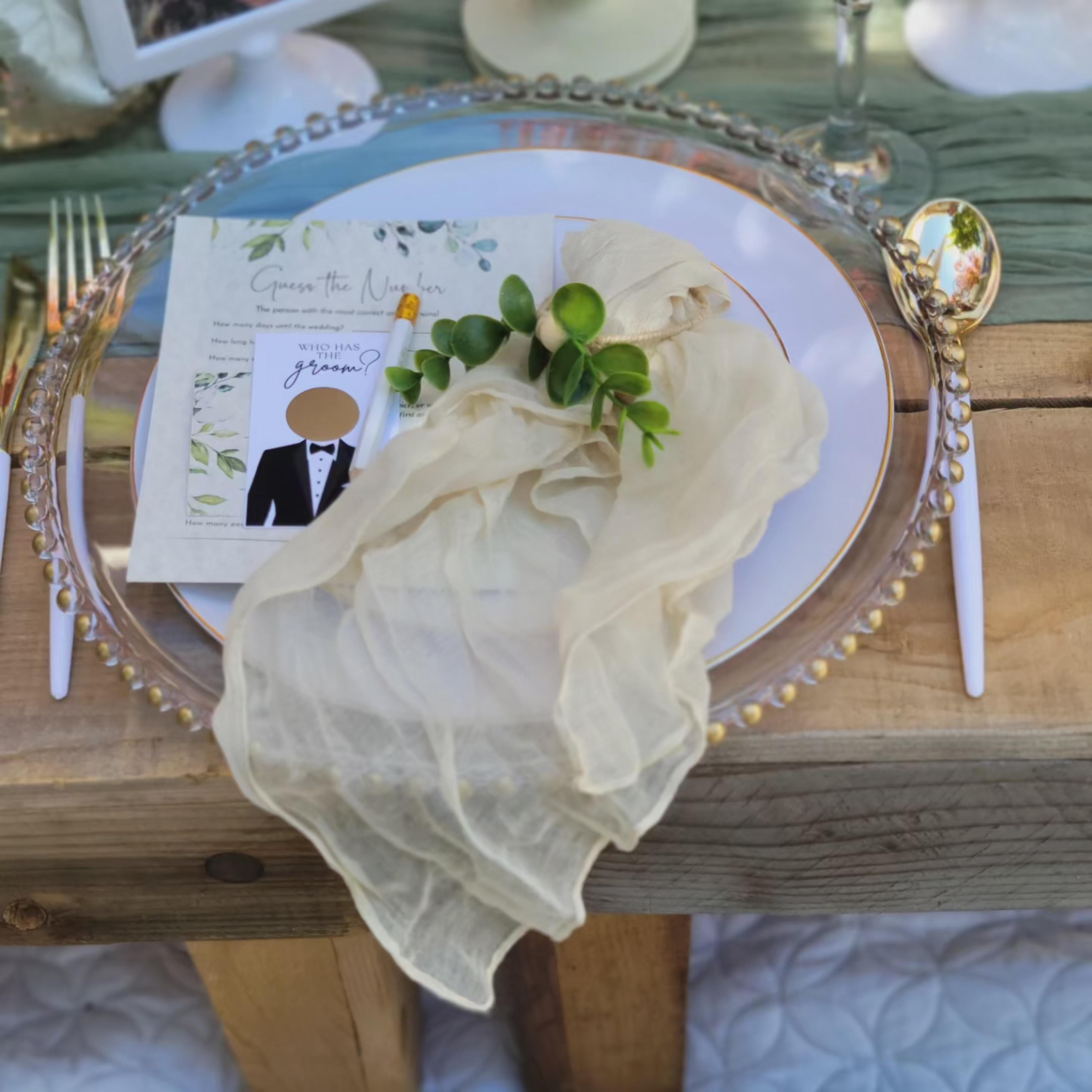 A elegantly set dining table features a white plate with a gold rim, adorned with a menu card and a delicate piece of fabric draped over greenery.