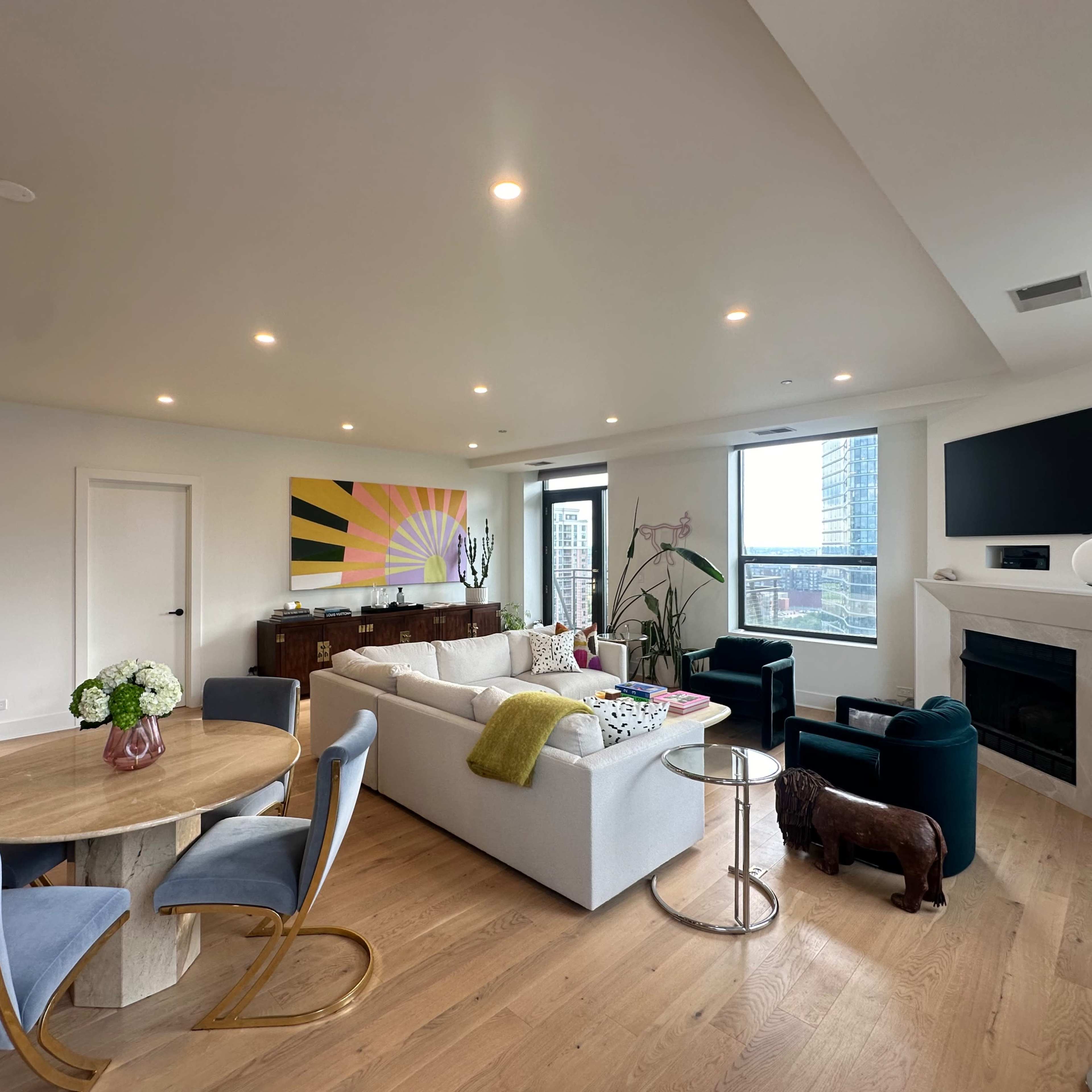 A modern living room with a dining area, featuring a round wooden table, a white sectional sofa, decorative artwork, and large windows offering a city view.
