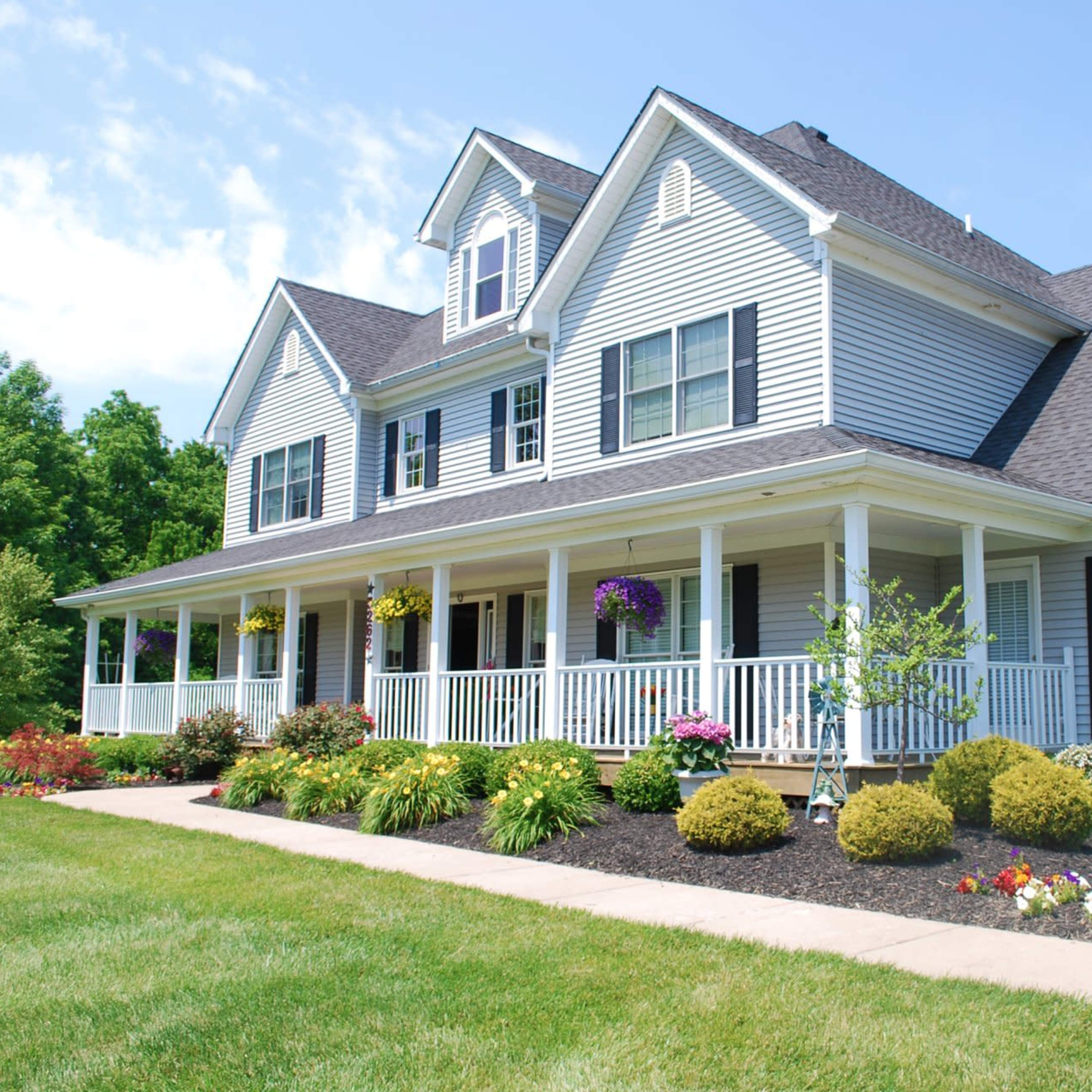 A large, two-story suburban house with a covered front porch and landscaped garden featuring colorful flowers and shrubs.