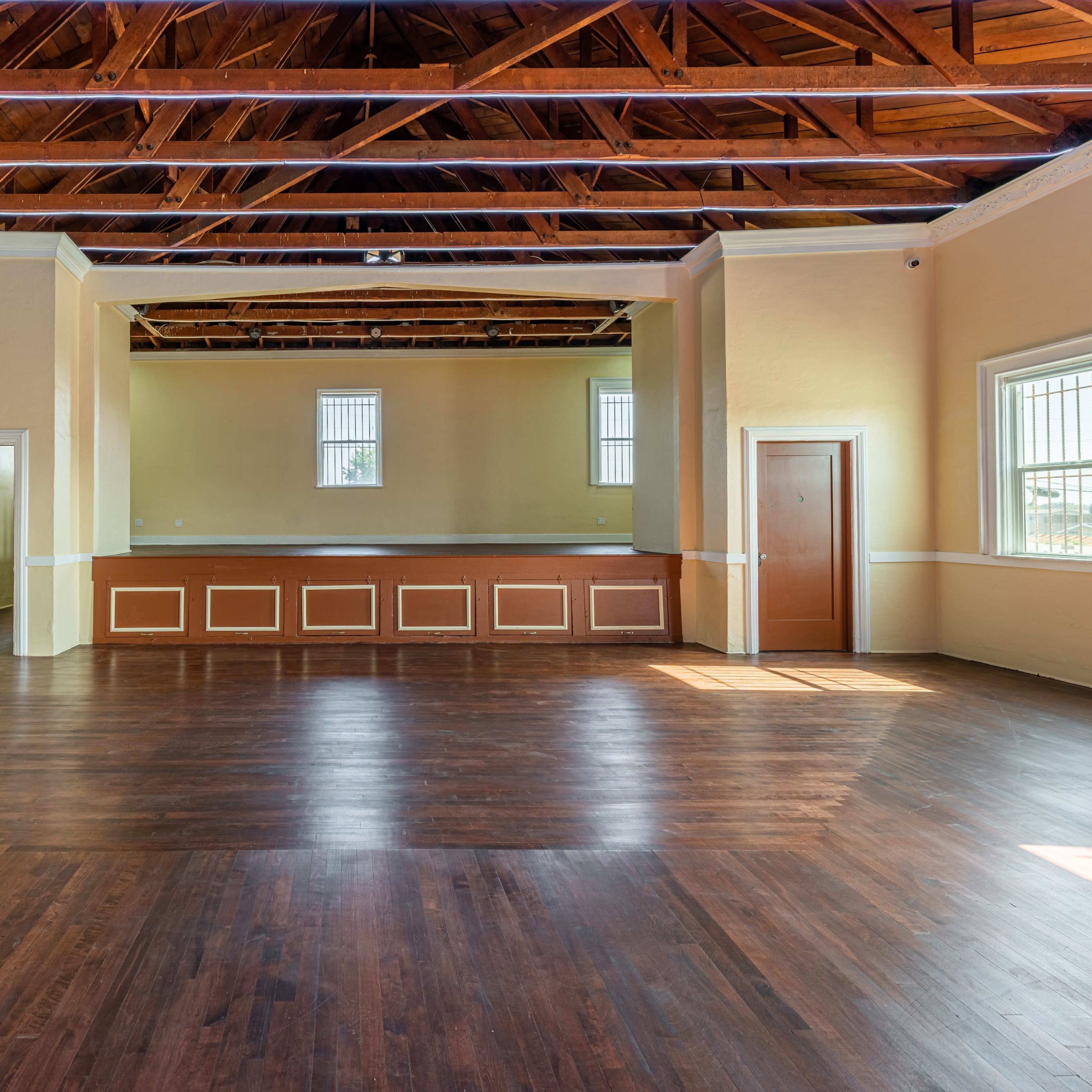 A spacious interior room with wooden flooring and high ceilings, featuring large windows and a raised platform at one end.