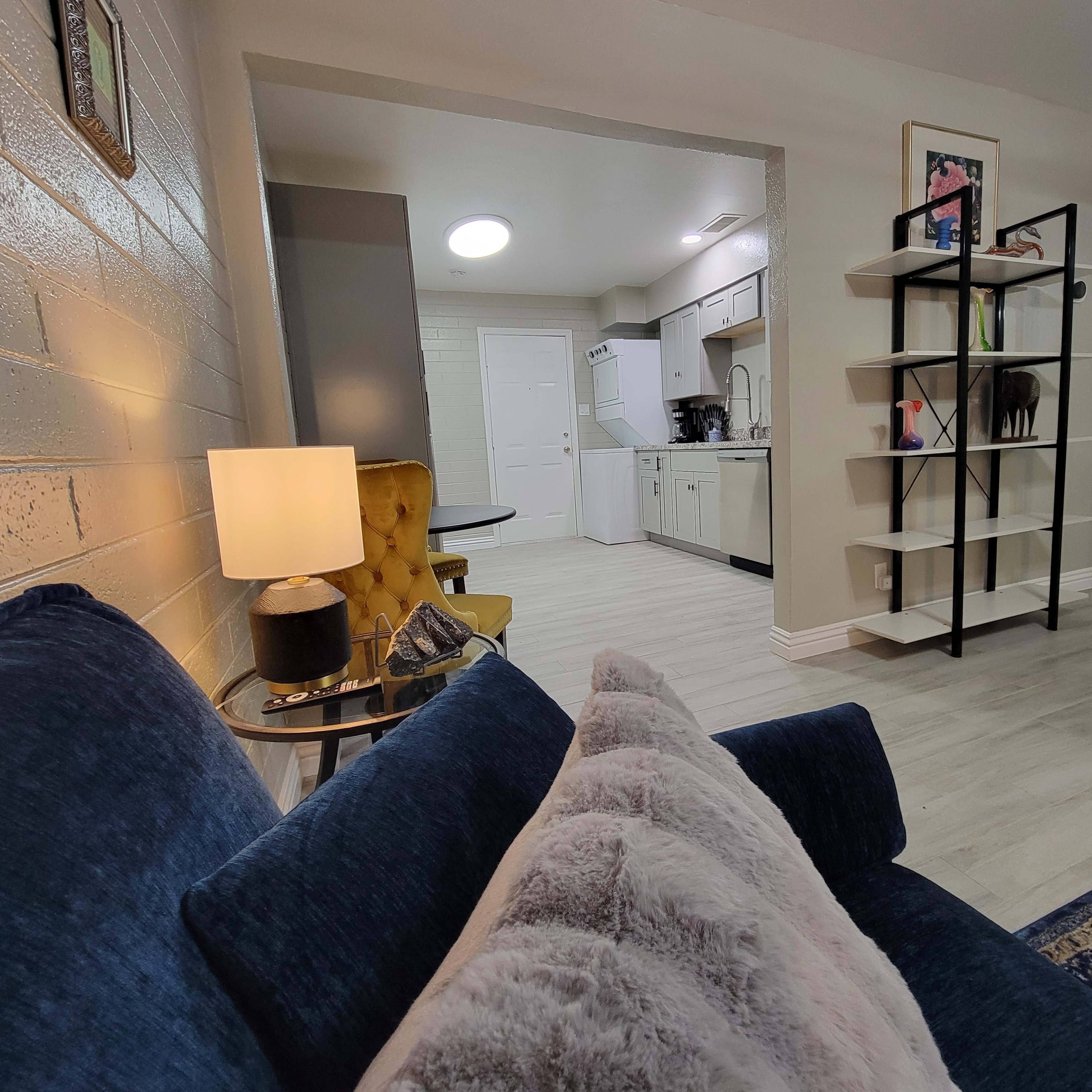 A cozy living room area with a blue couch, a lamp, and a bookshelf, leading into an open kitchen space.