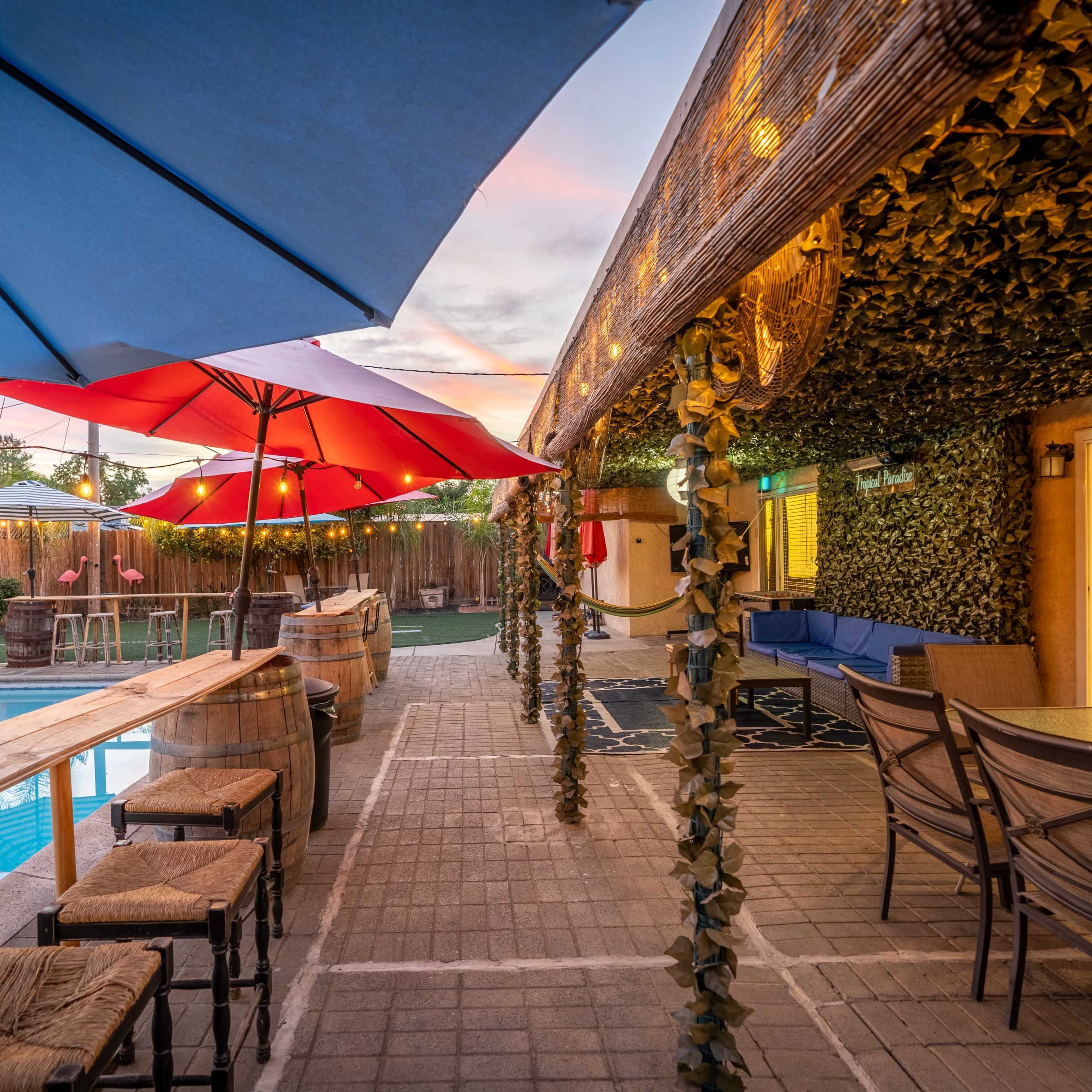 A swimming pool area with a bar, blue and red umbrellas, and seating surrounded by greenery and string lights.