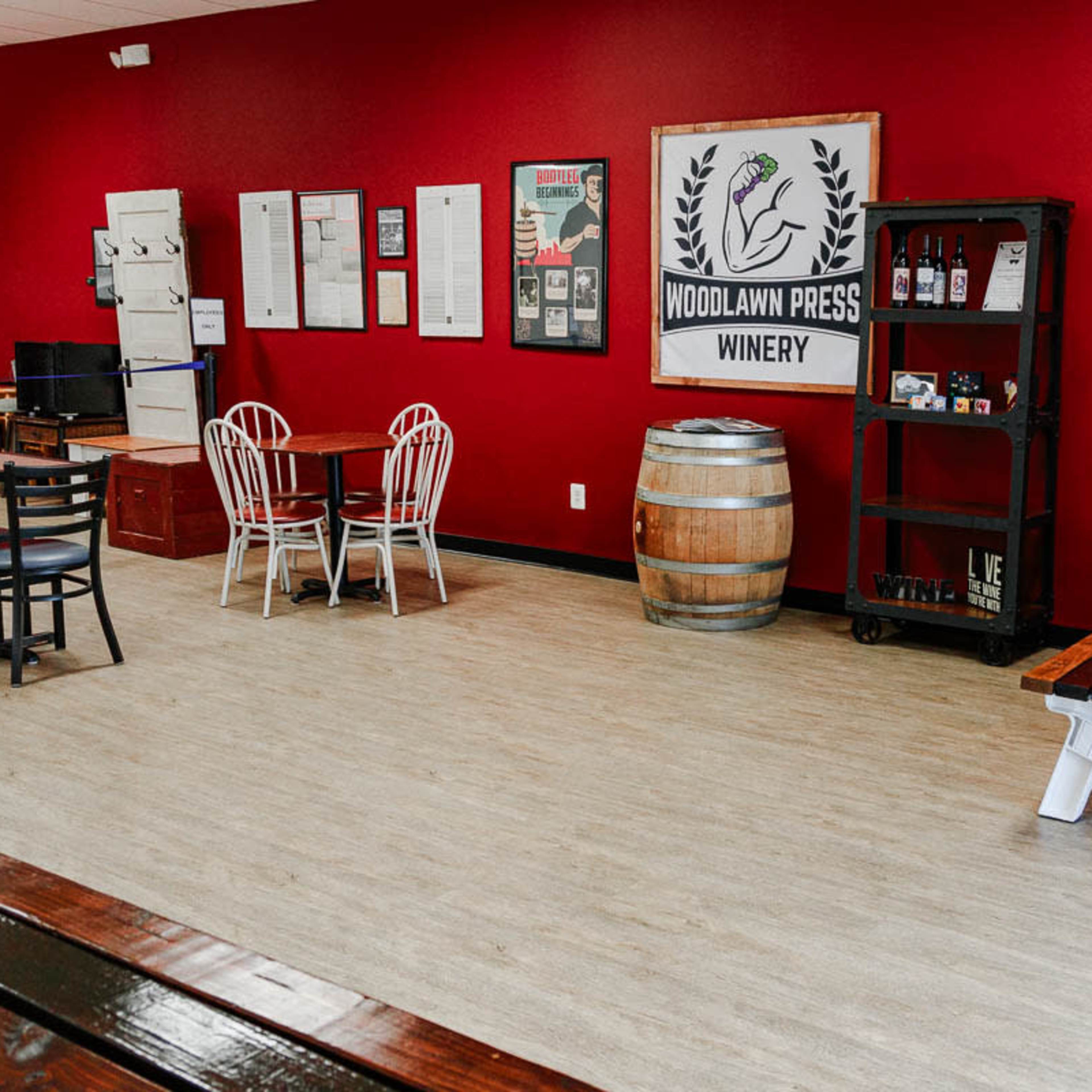 The interior of a winery features red walls, wooden tables, and a barrel, along with informational posters and a sign for "Woodlawn Press Winery."