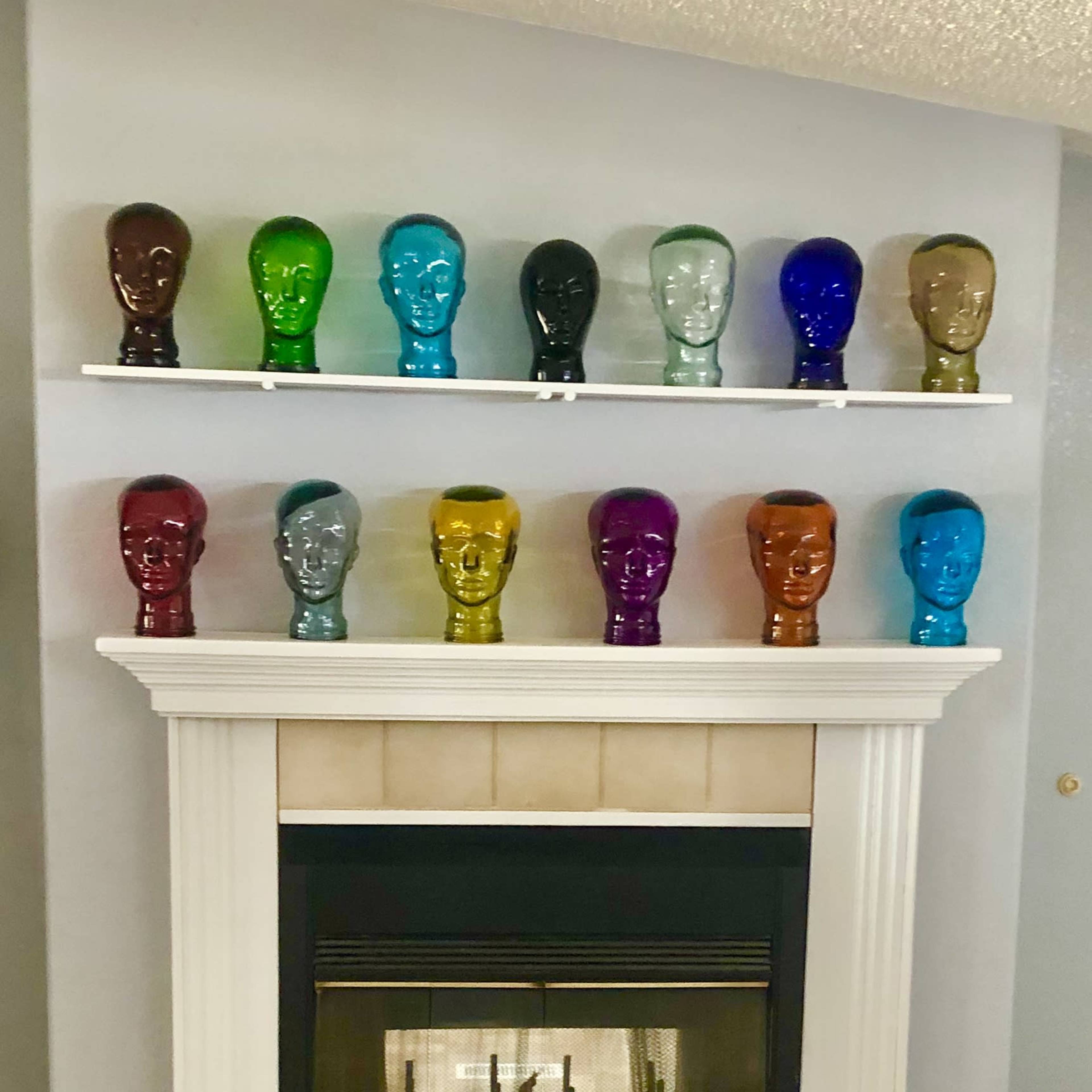 A row of colorful glass head sculptures is displayed on a shelf above a white fireplace in a room with gray walls.