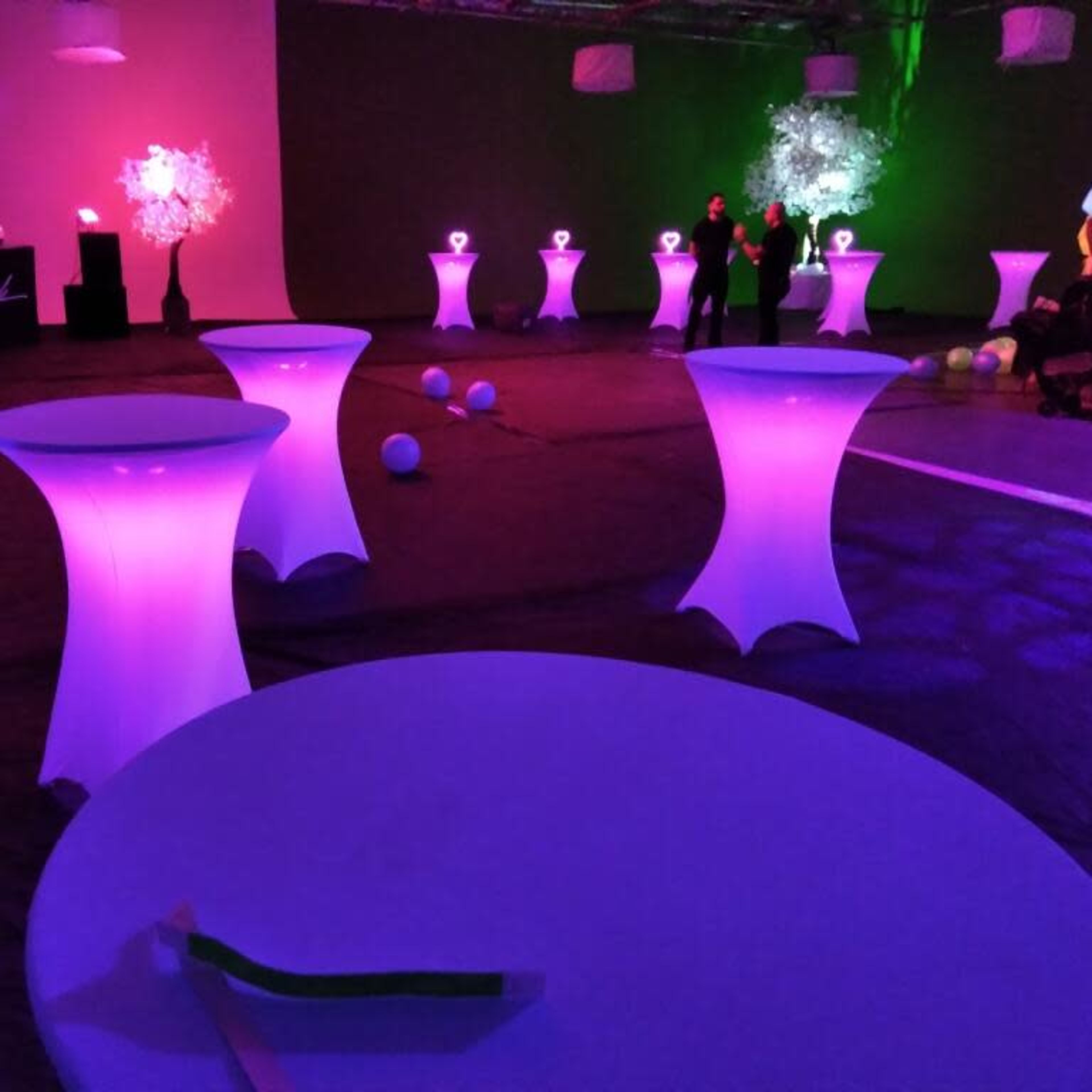 A dimly lit event space with illuminated tables, decorated trees, and a bar area in the background.