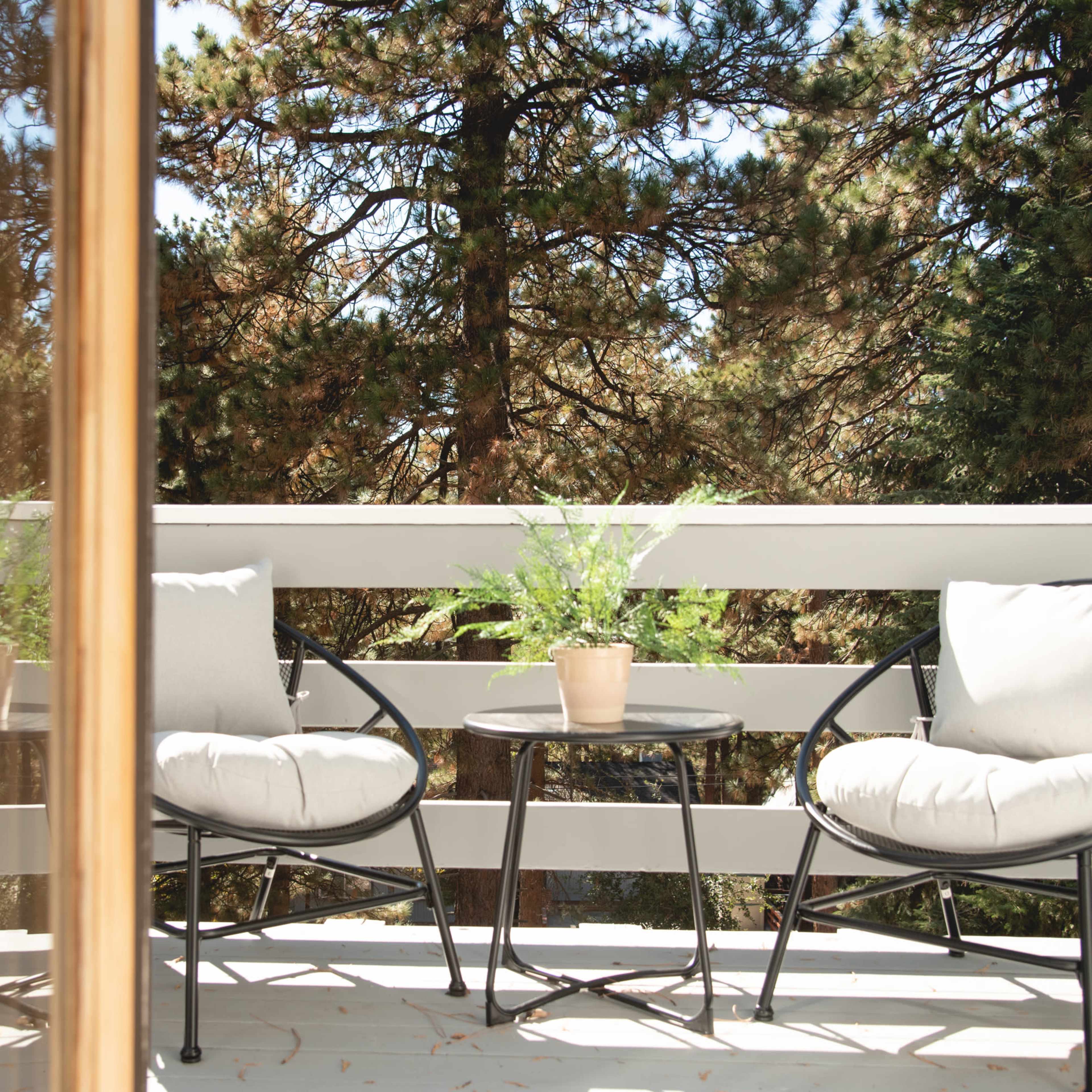 A small outdoor balcony features two black chairs and a table with a potted plant, surrounded by tall trees.