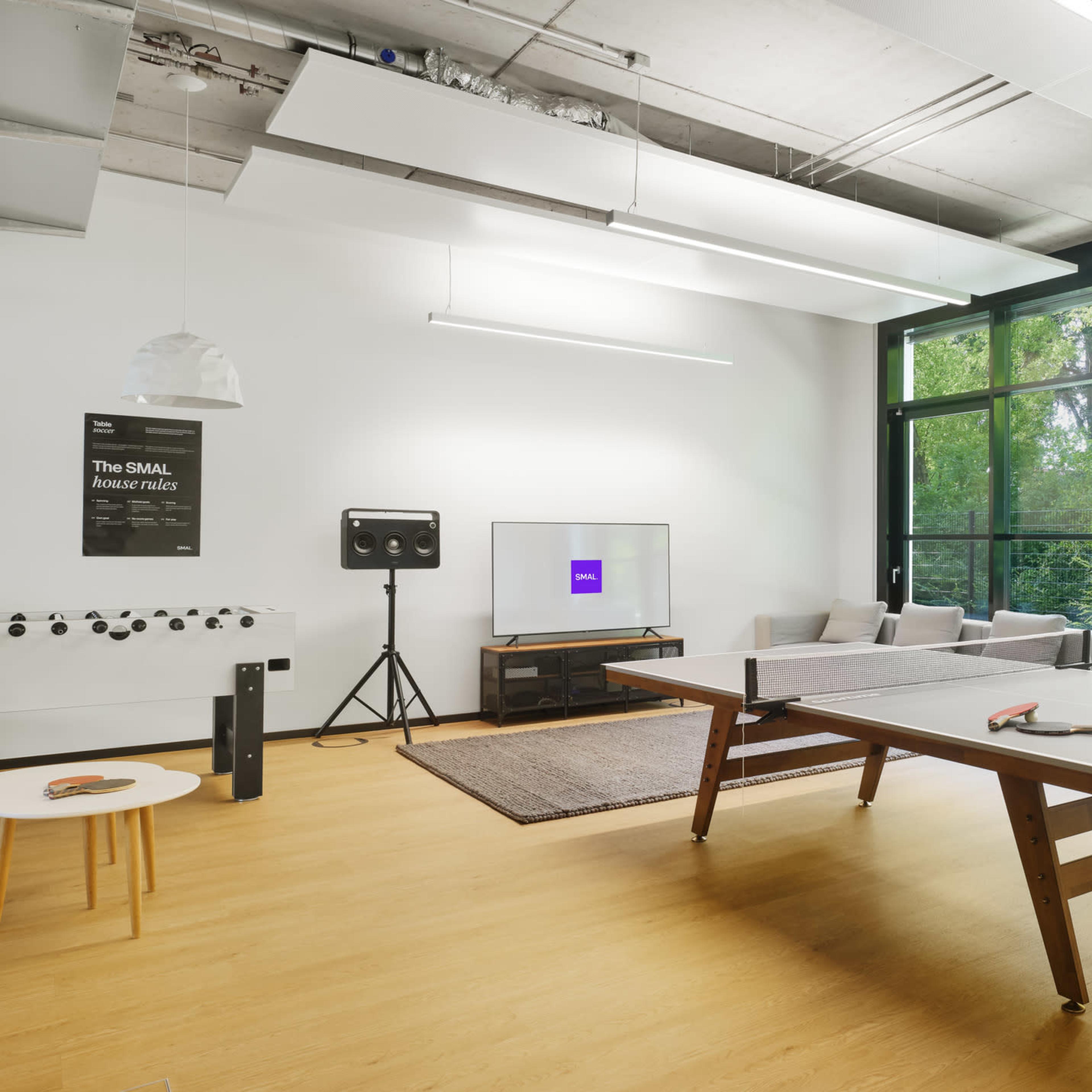 A modern, well-lit room features a ping-pong table, a television on a stand, and seating arranged near large windows overlooking greenery.