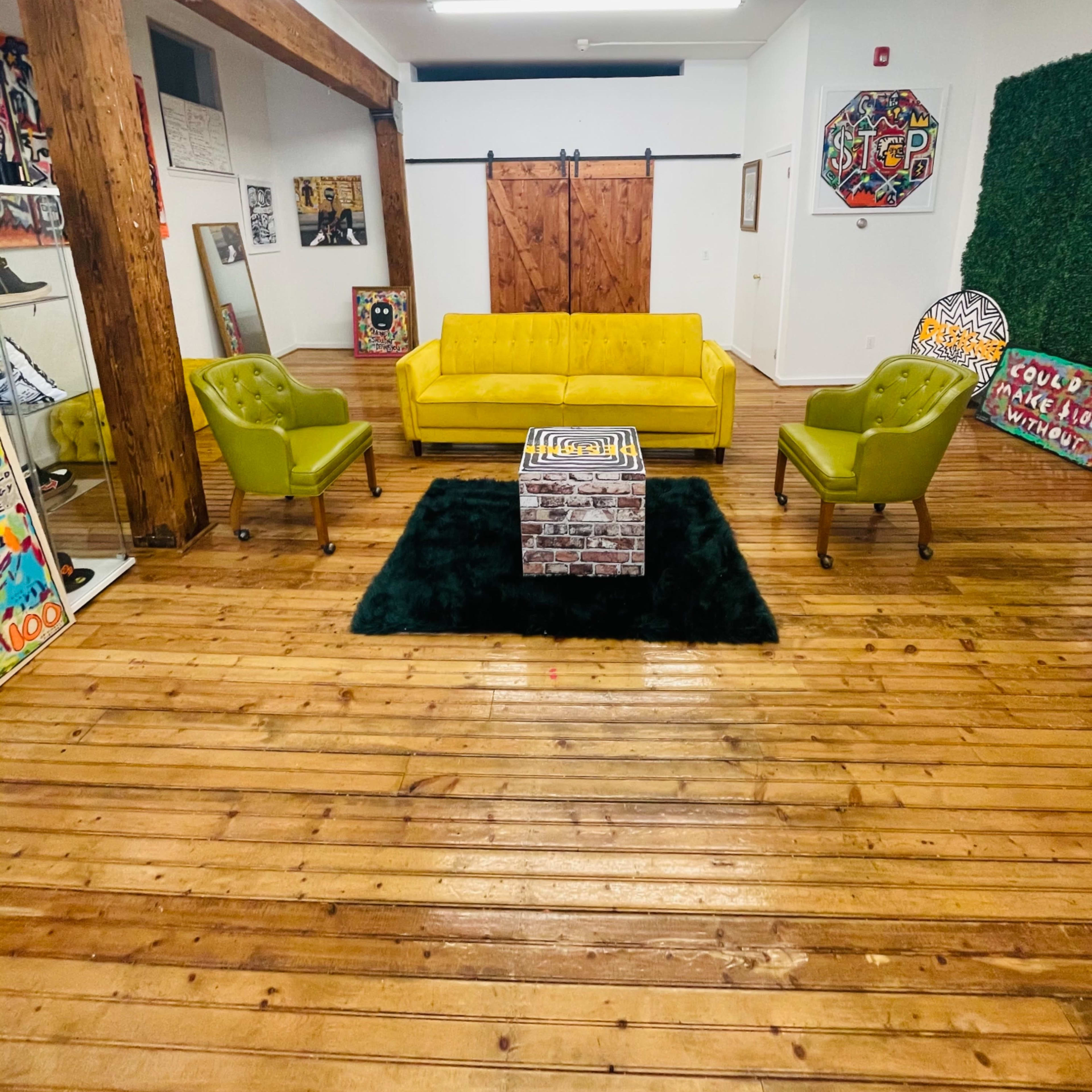 A bright room features a yellow sofa, two green armchairs, and a square coffee table on a dark rug, with wooden floors and exposed beams.