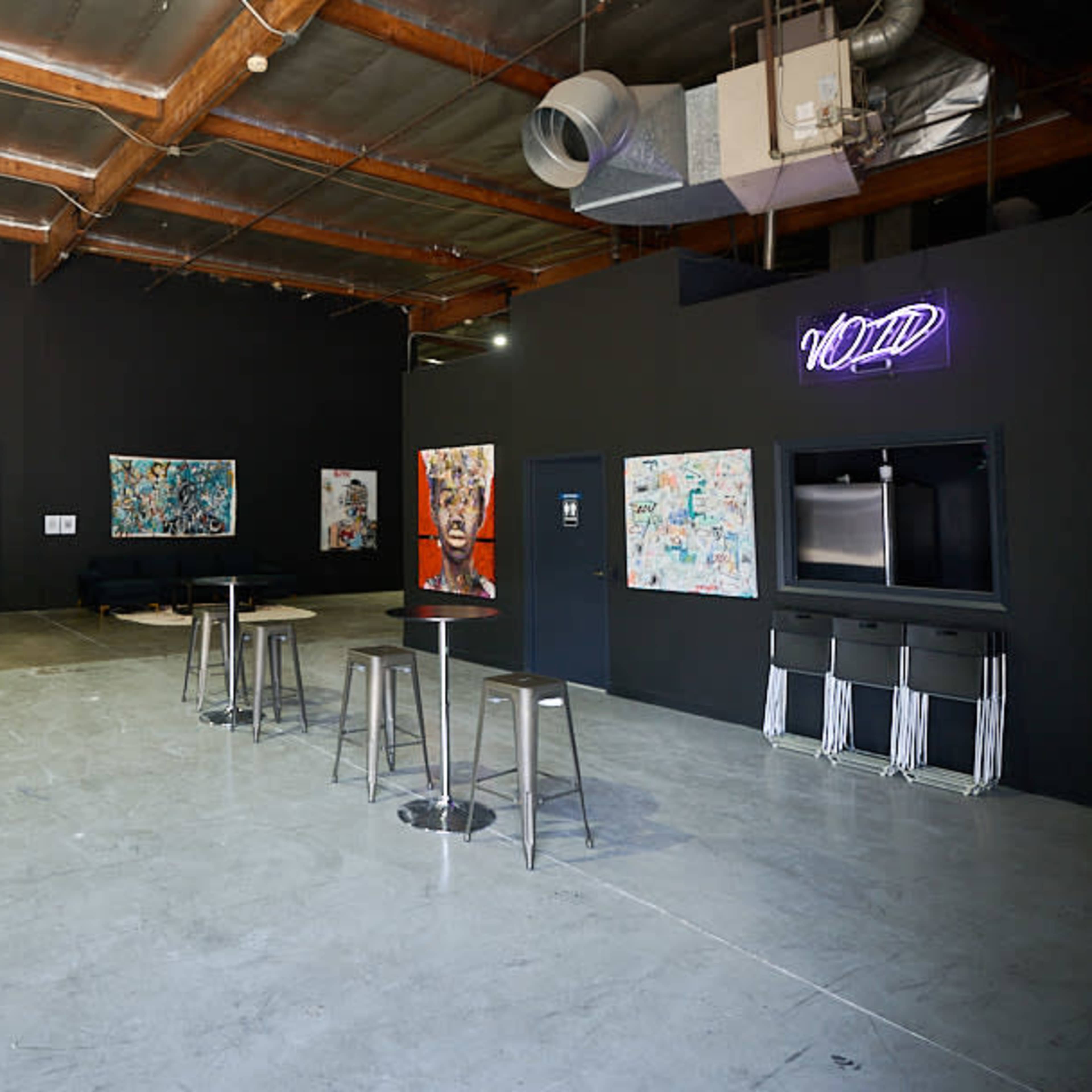 An interior space with dark walls, several abstract artworks displayed, and bar stools arranged around a small table.