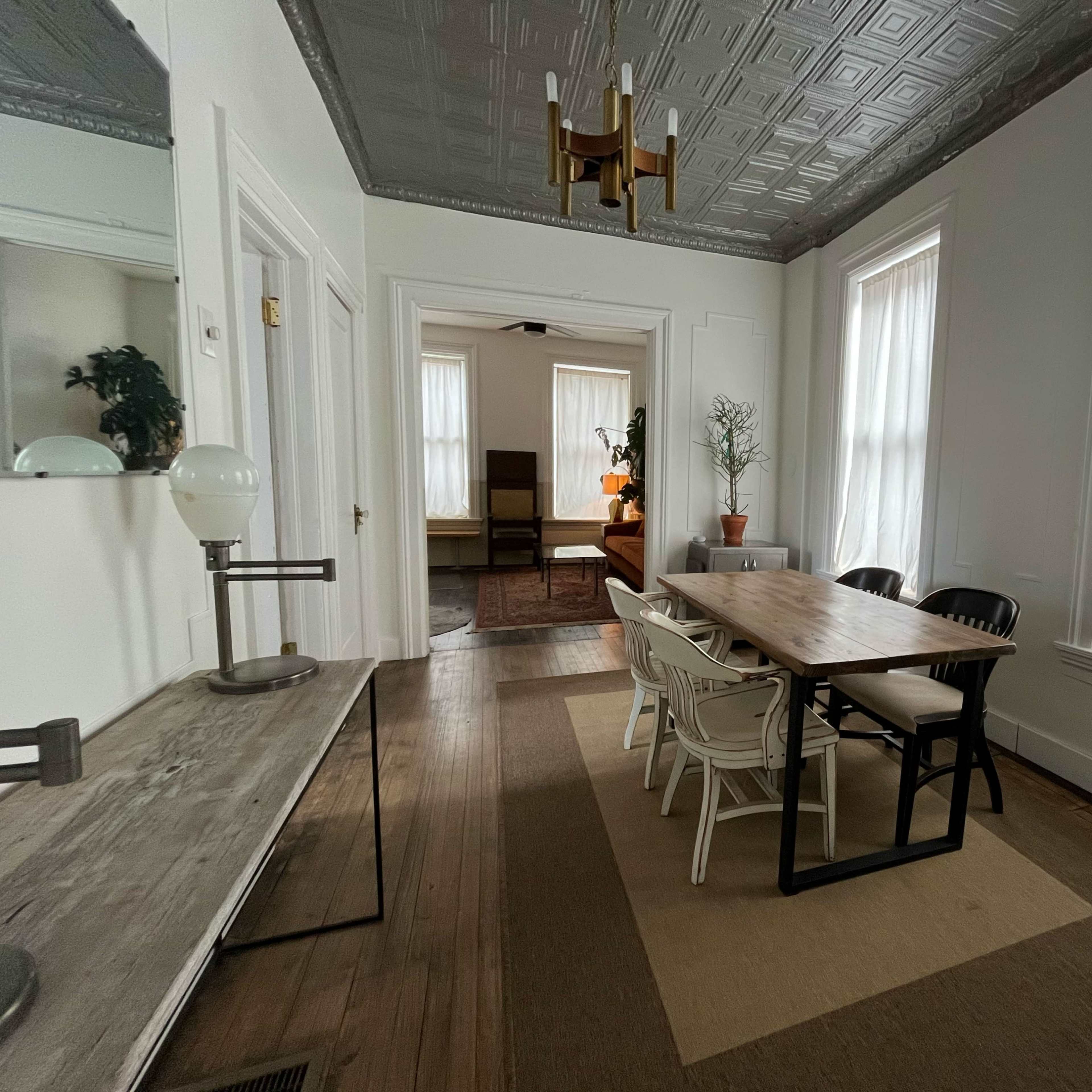 A dining area with a long wooden table surrounded by chairs, a side console table, and large windows with sheer curtains.