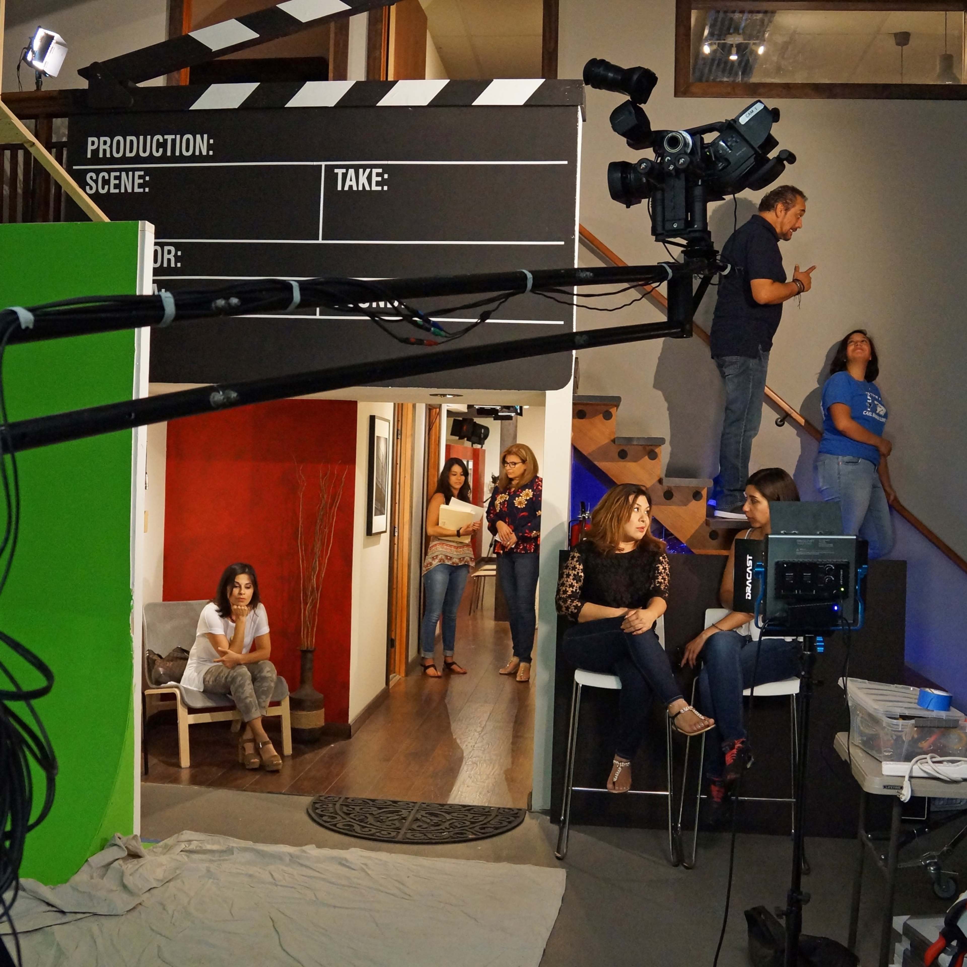 A film set features a green screen, a camera mounted on a crane, and crew members preparing for a shoot, while several people sit in the background.