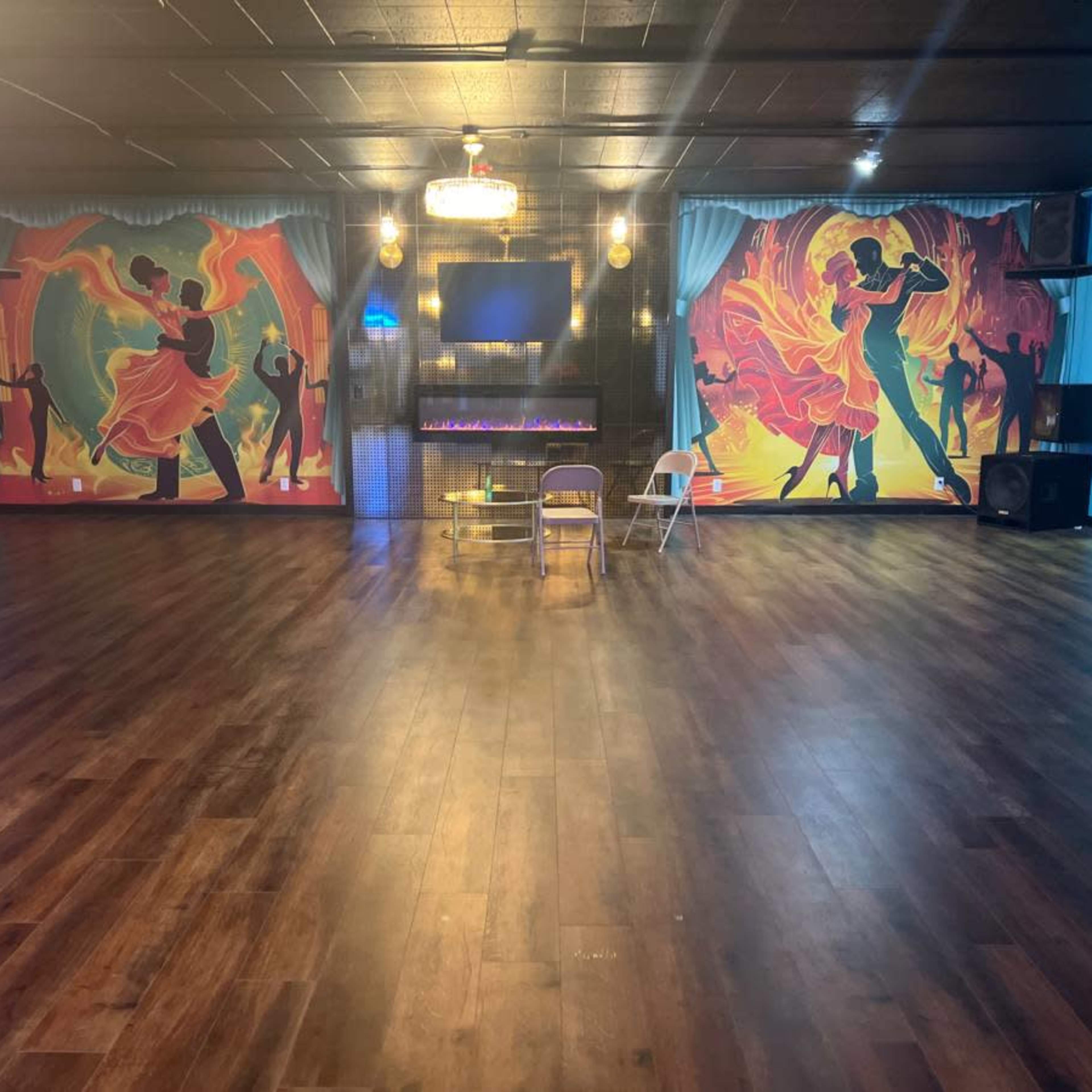 A spacious dance studio with colorful murals on the walls, a large wooden floor, and a modern electric fireplace.