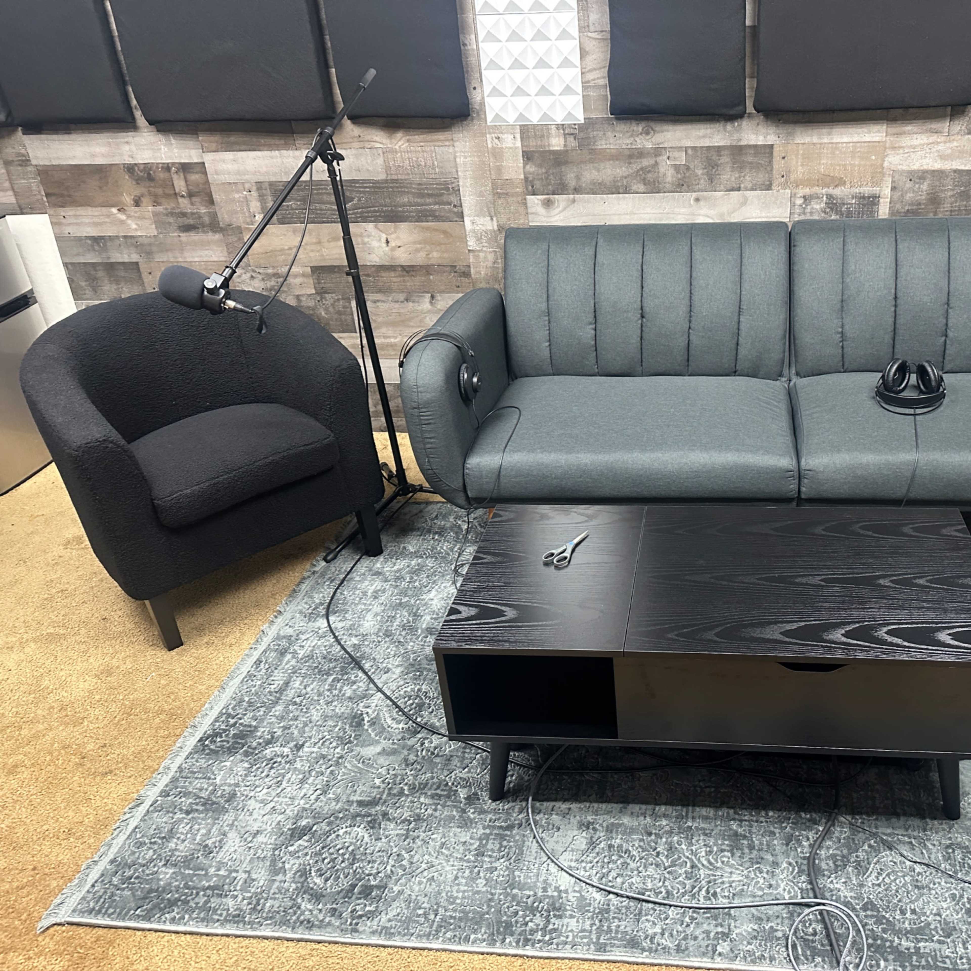 A gray couch and a black chair positioned around a black coffee table on a patterned rug in a studio-like setting.