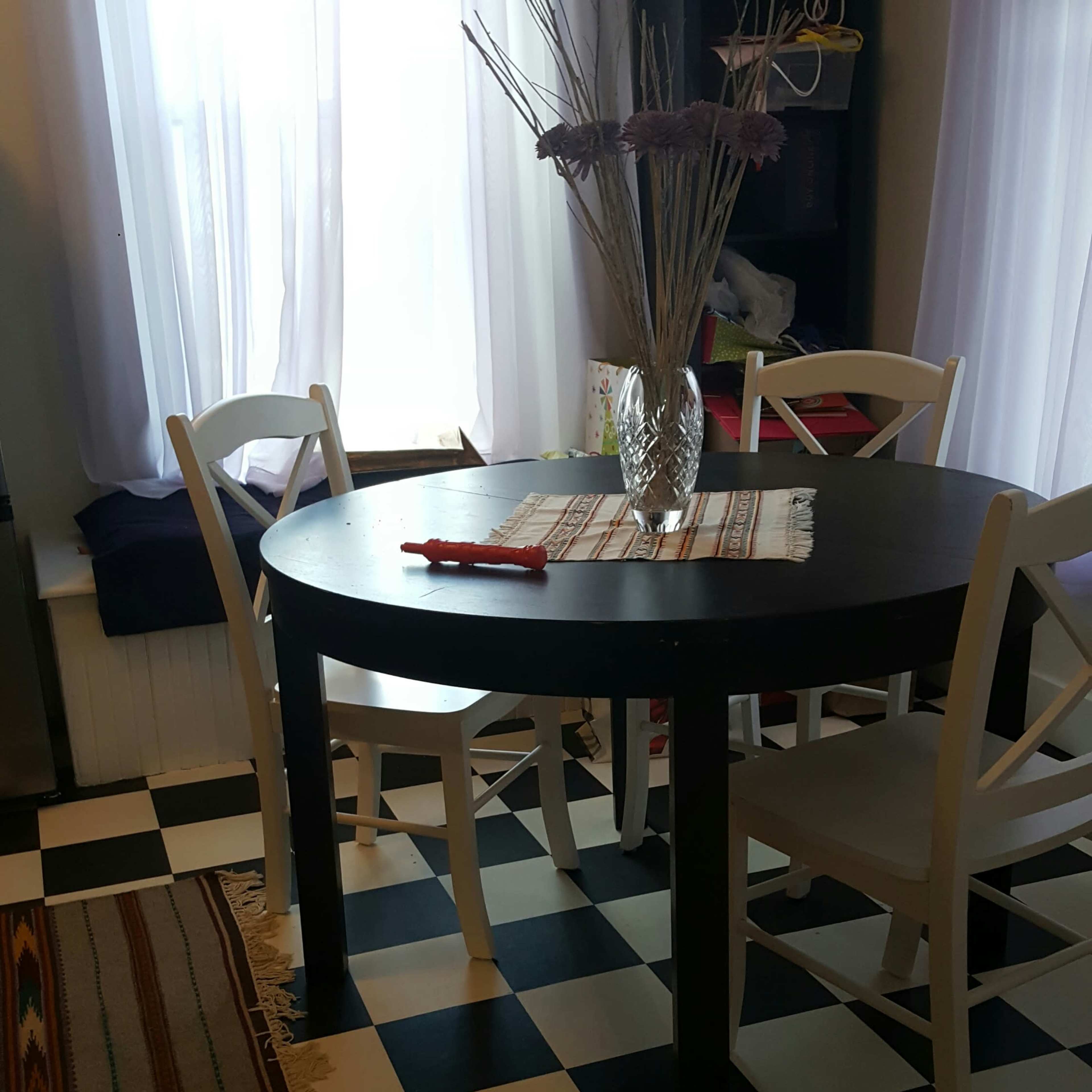 A round black table sits in a room with checkered floors, surrounded by white chairs and decorated with a vase of flowers.