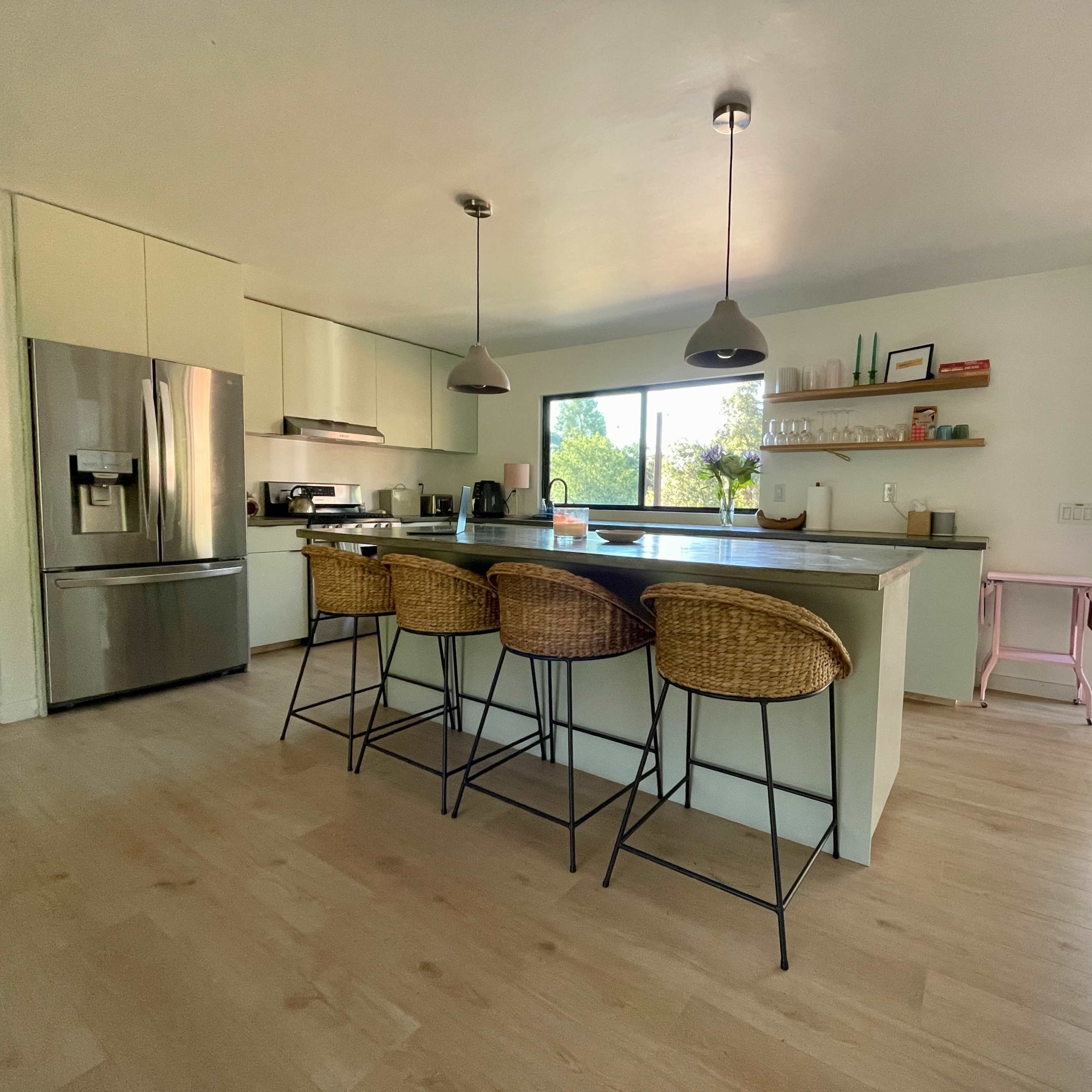 A modern kitchen features a large island with four bar stools, stainless steel appliances, and a sliding door opening to an outdoor deck.