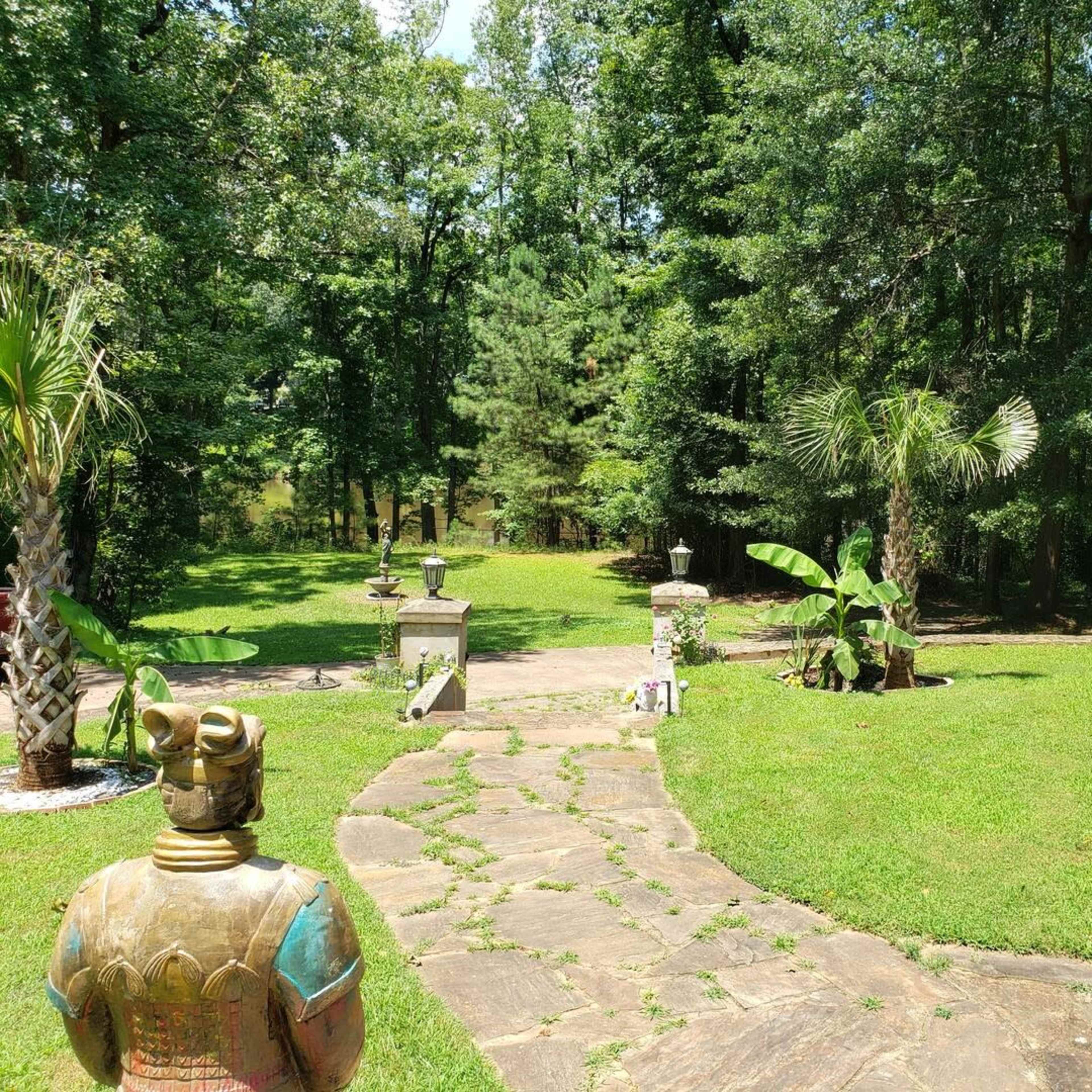 A landscaped garden with stone pathways, sculptures, and lush greenery surrounded by trees.