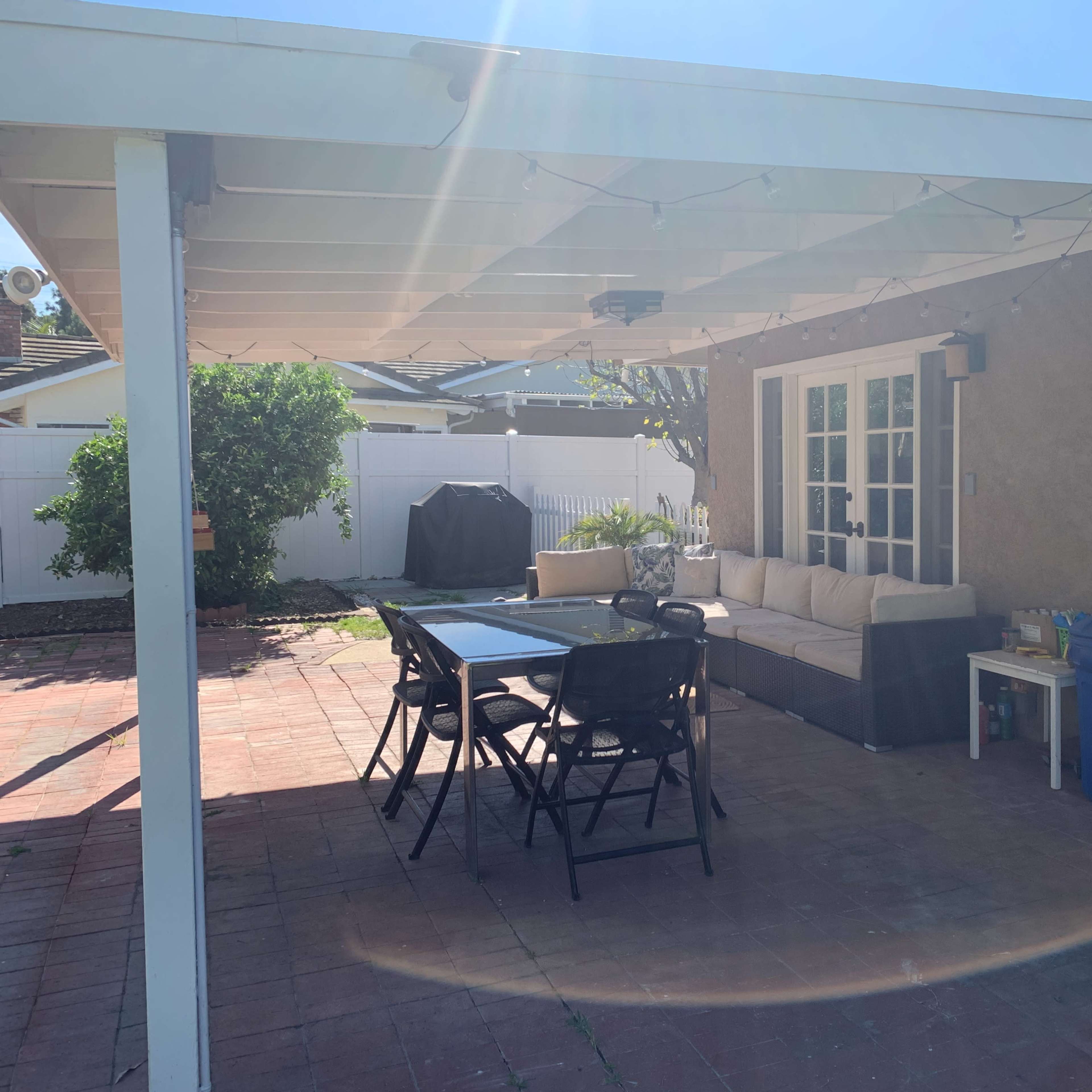 A patio area with a dining table, chairs, a sofa, and a blue trash can, set against a backdrop of a house and a fenced yard.