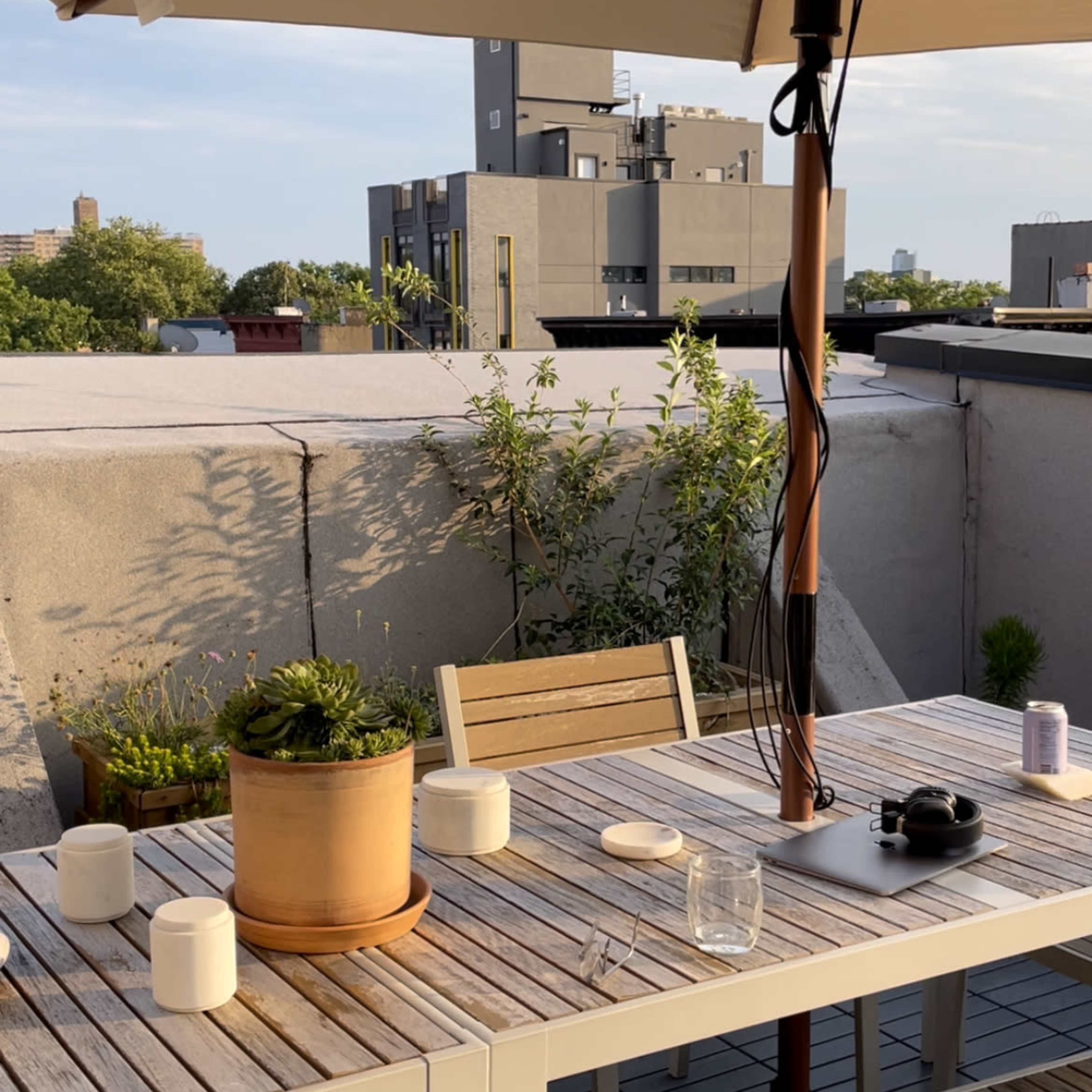 A rooftop patio features a wooden table with potted plants, candles, and an umbrella against a backdrop of buildings.