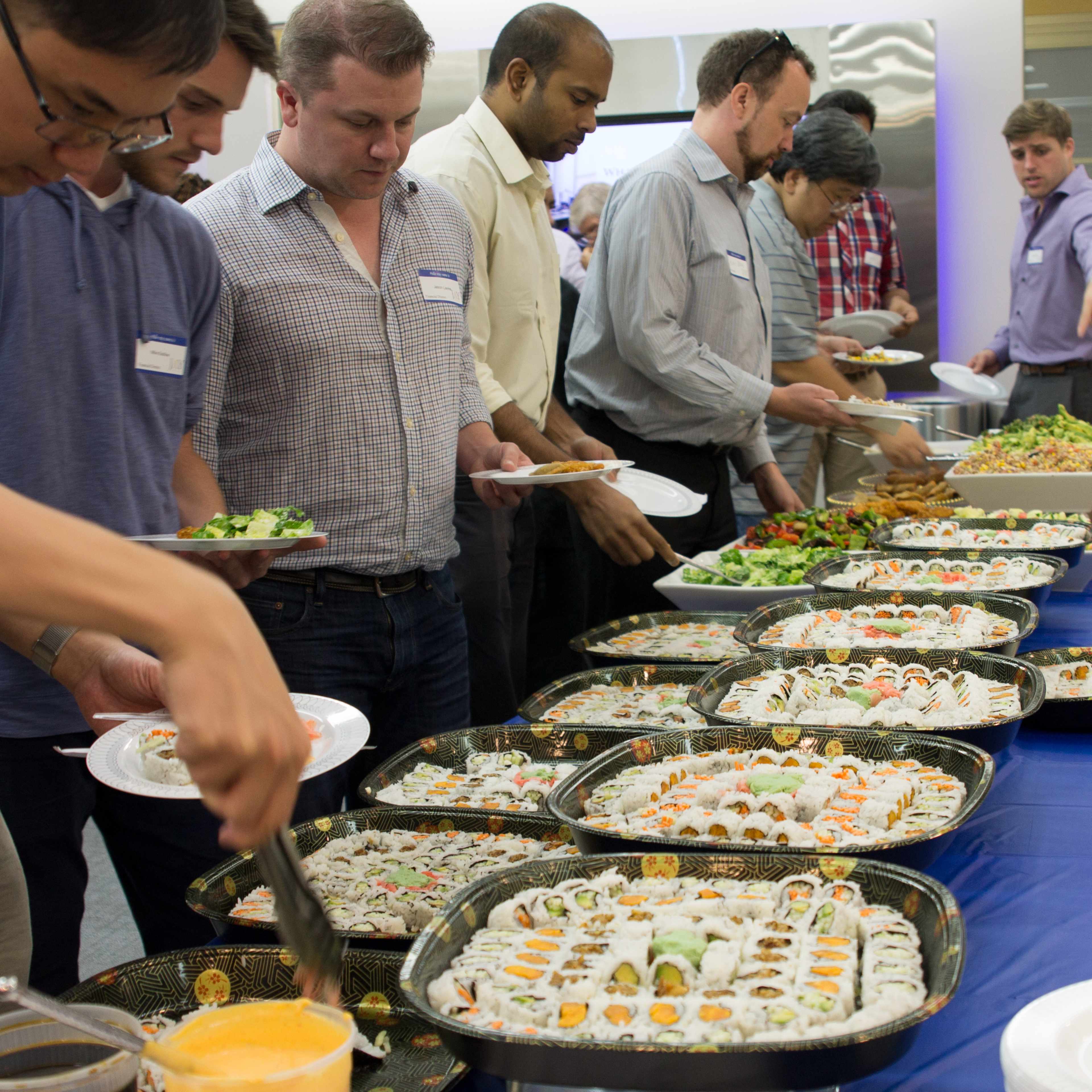 A group of people is serving themselves food from a buffet table filled with various sushi dishes and salads.